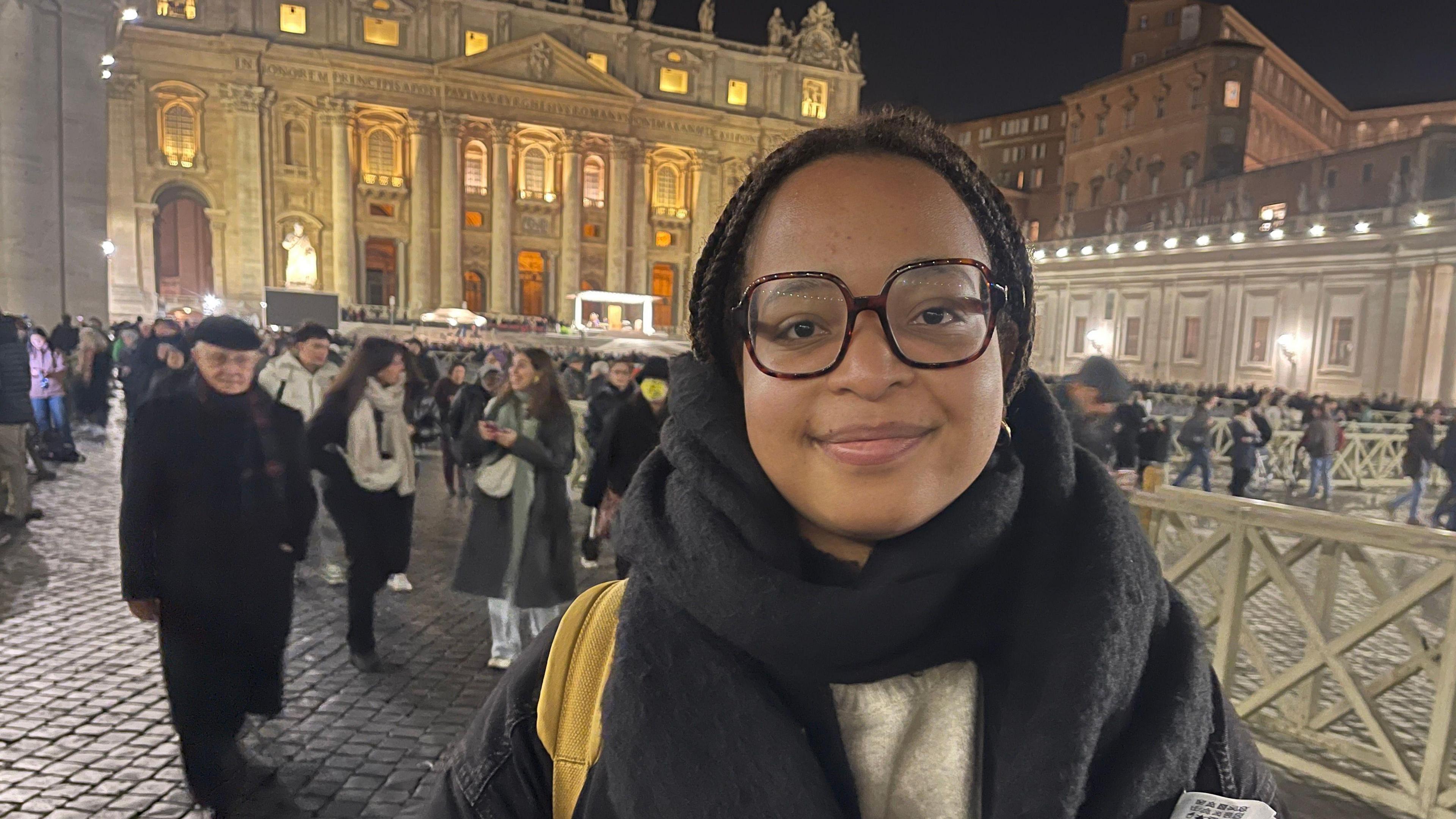 Stacey, a medical student from Paris, is a young woman, with large statement glasses, a scarf, a cream jumper, and has a visible yellow strap of a backpack. She is looking directly at the camera, with crowds and the basilica visible behind her