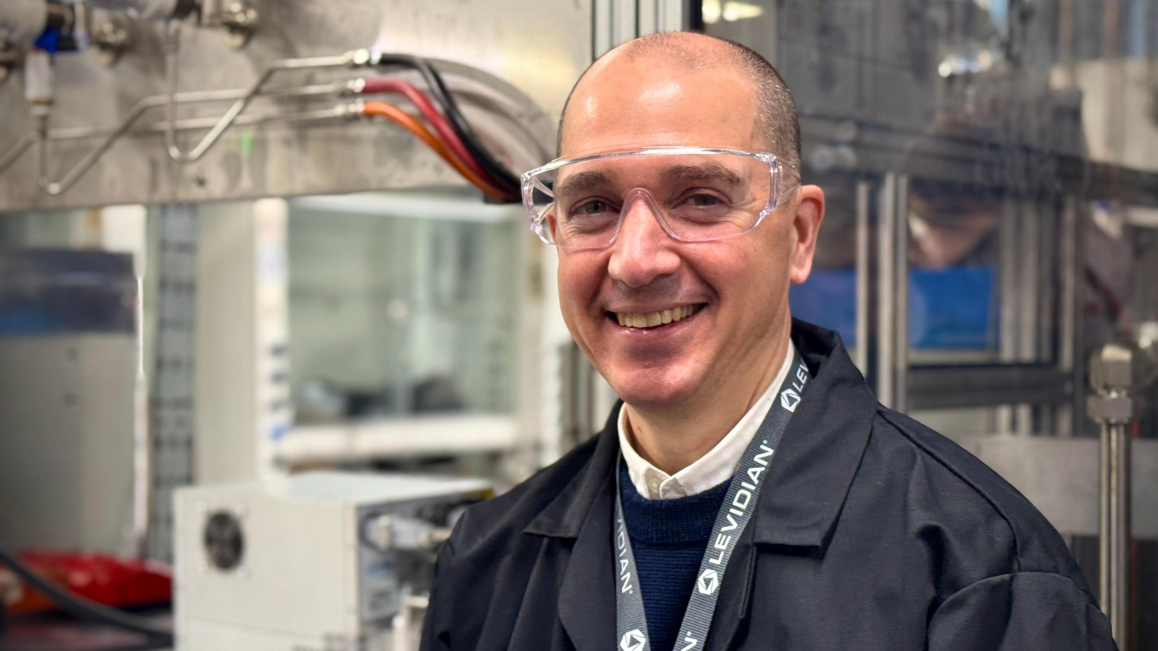 Levidian Chief Technology Officer Alistair Donaldson wearing a lab coat and goggles. He is standing, smiling, alongside a small version of the Loop system in Cambridge