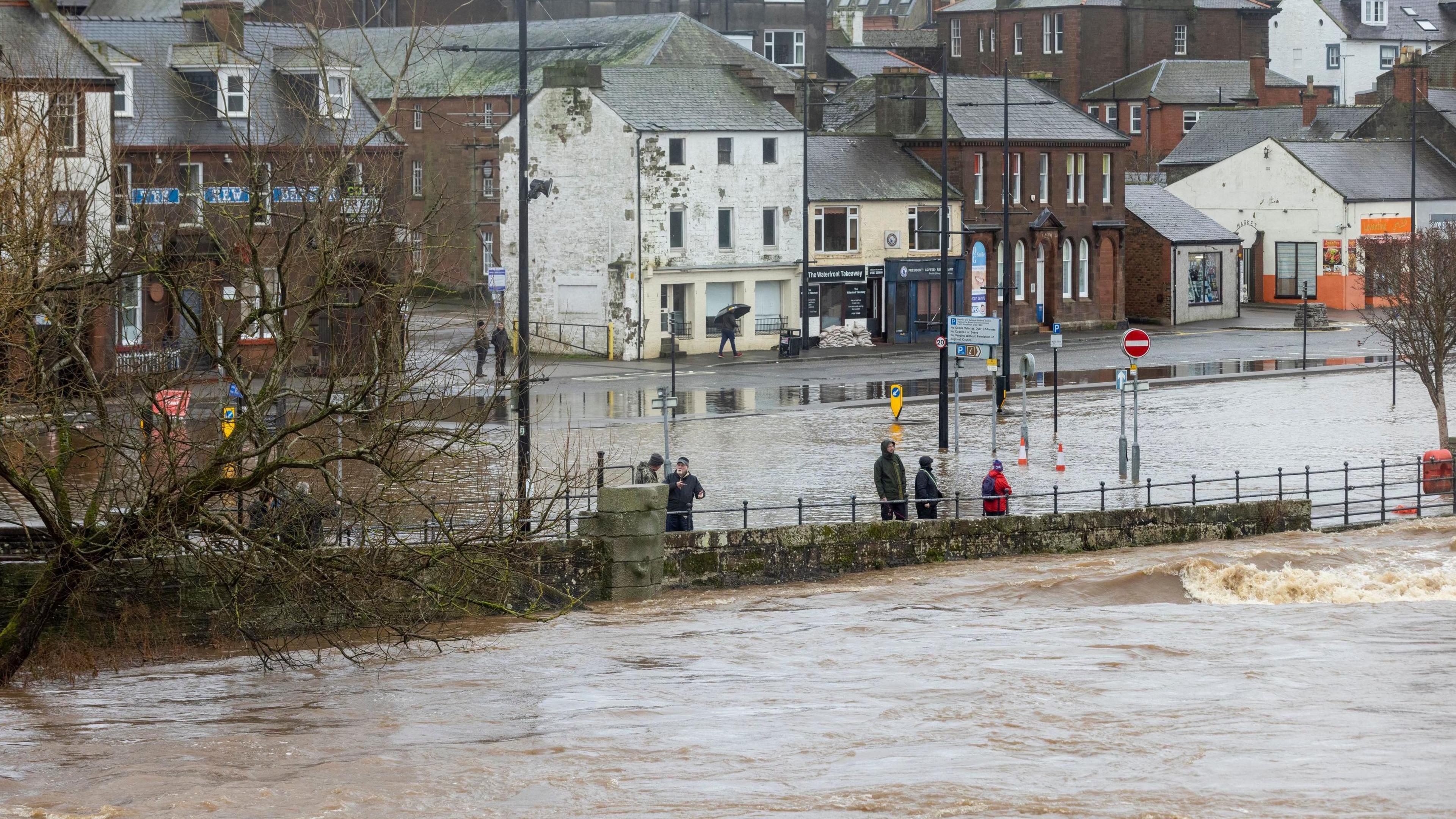 The Whitesands area of Dumfries is flooded by water from the River Nith moving up towards a row of businesses with a small number of people standing watching the scene