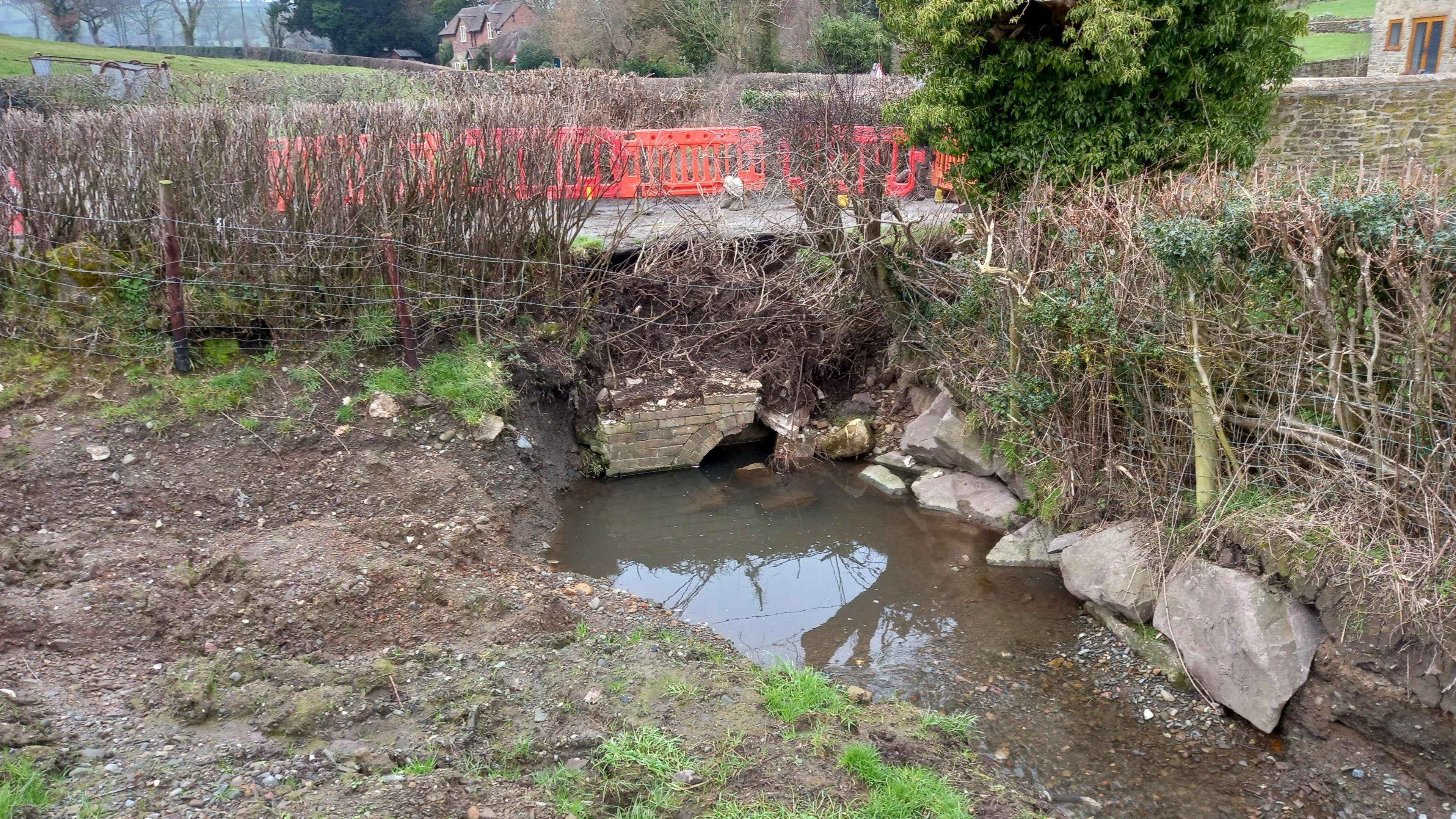 A broken culvert underneath a rural road. Water is visible at the mouth of the culvert with fenced-off field either side. Orange road barriers can be seen on the road above the culvert.