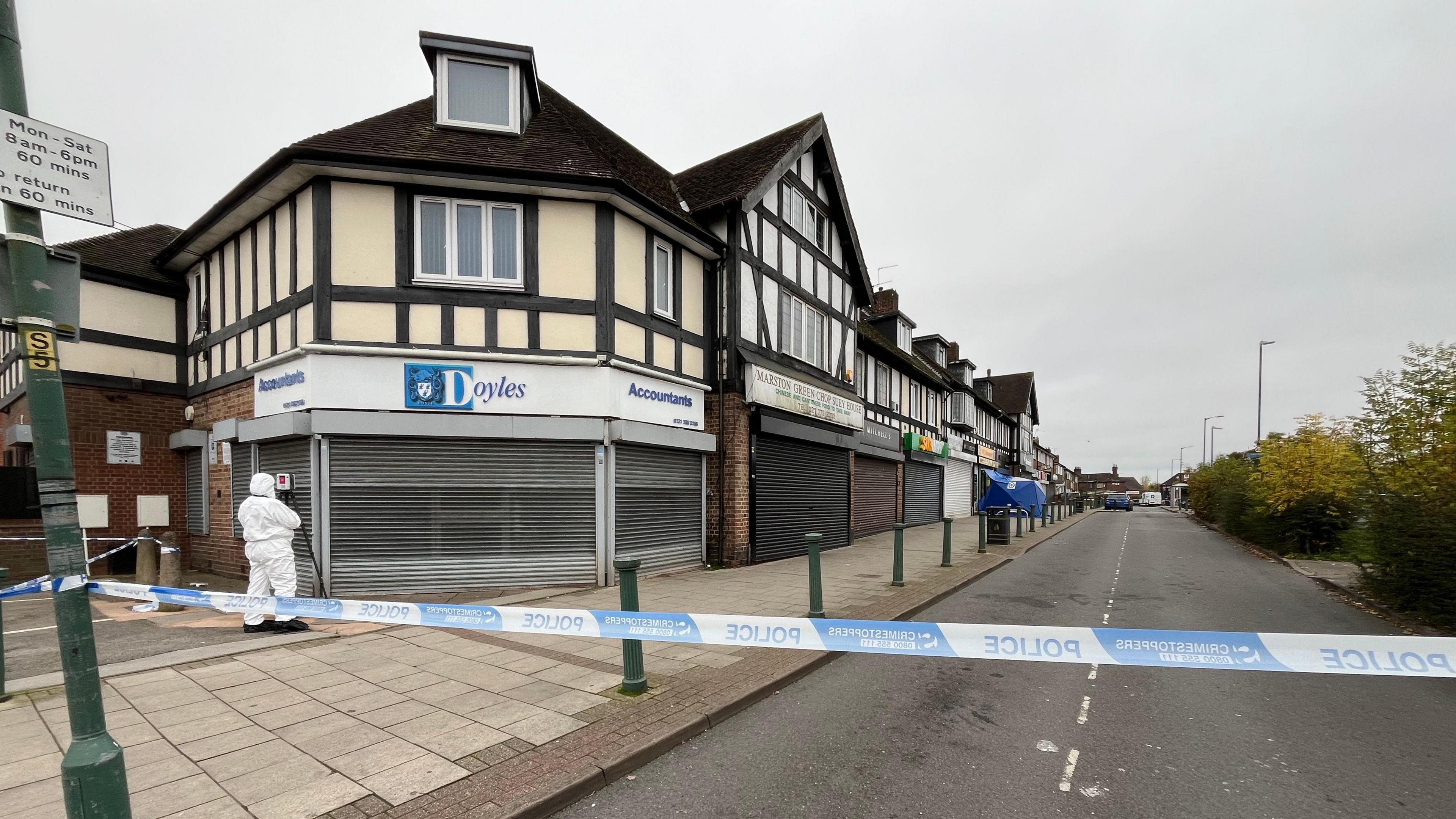 A road is cordoned off with blue and white police tape. There is a forensics worker in a white protective suit holding a camera with a tripod. In the distance, the blue forensics tent in front of a shop is visible.