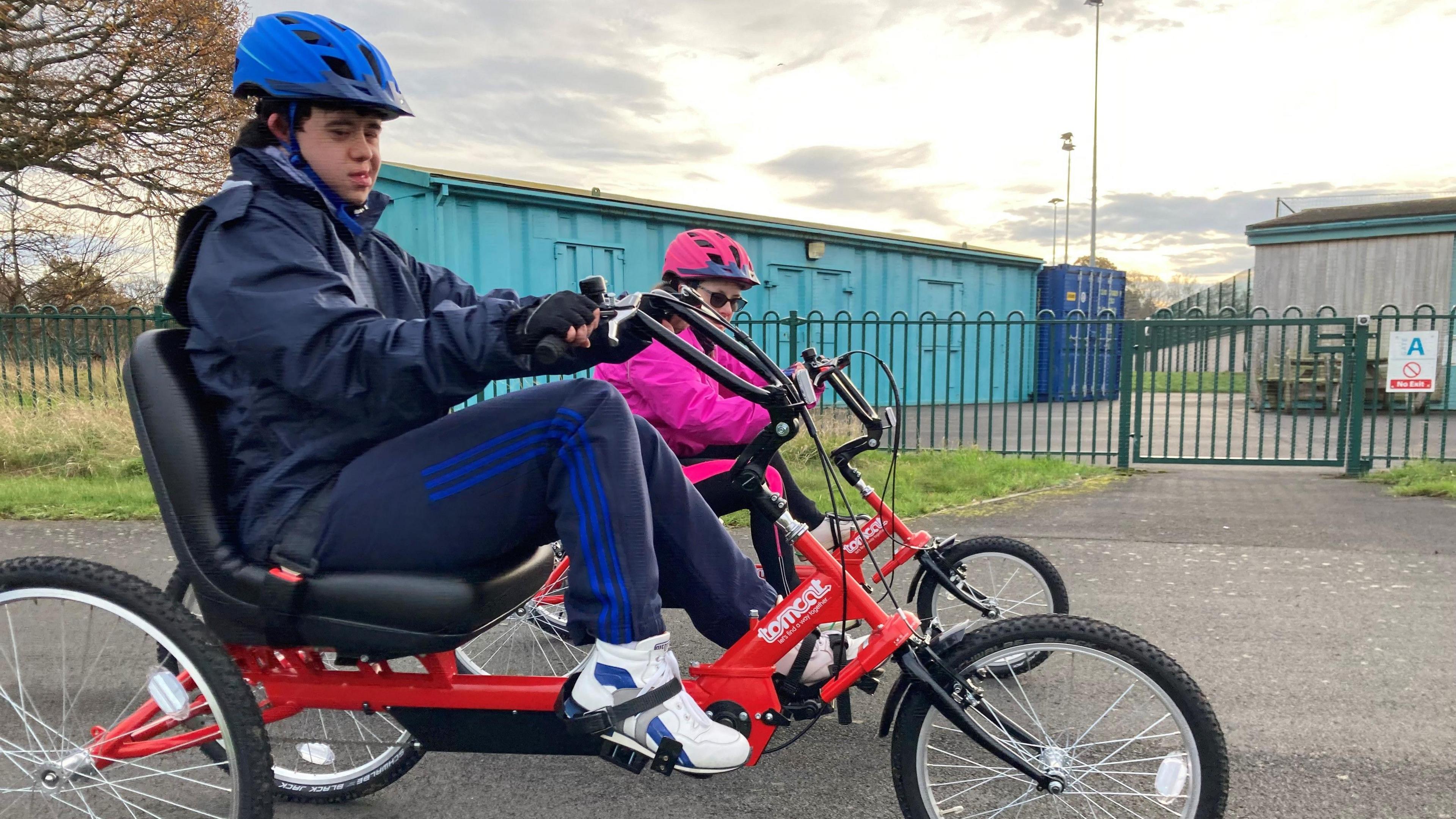 Two people are sitting on specially modified bicycles. One man in blue is wearing a helmet and smiling at the camera. Behind him, a lady is wearing a pink helmet.