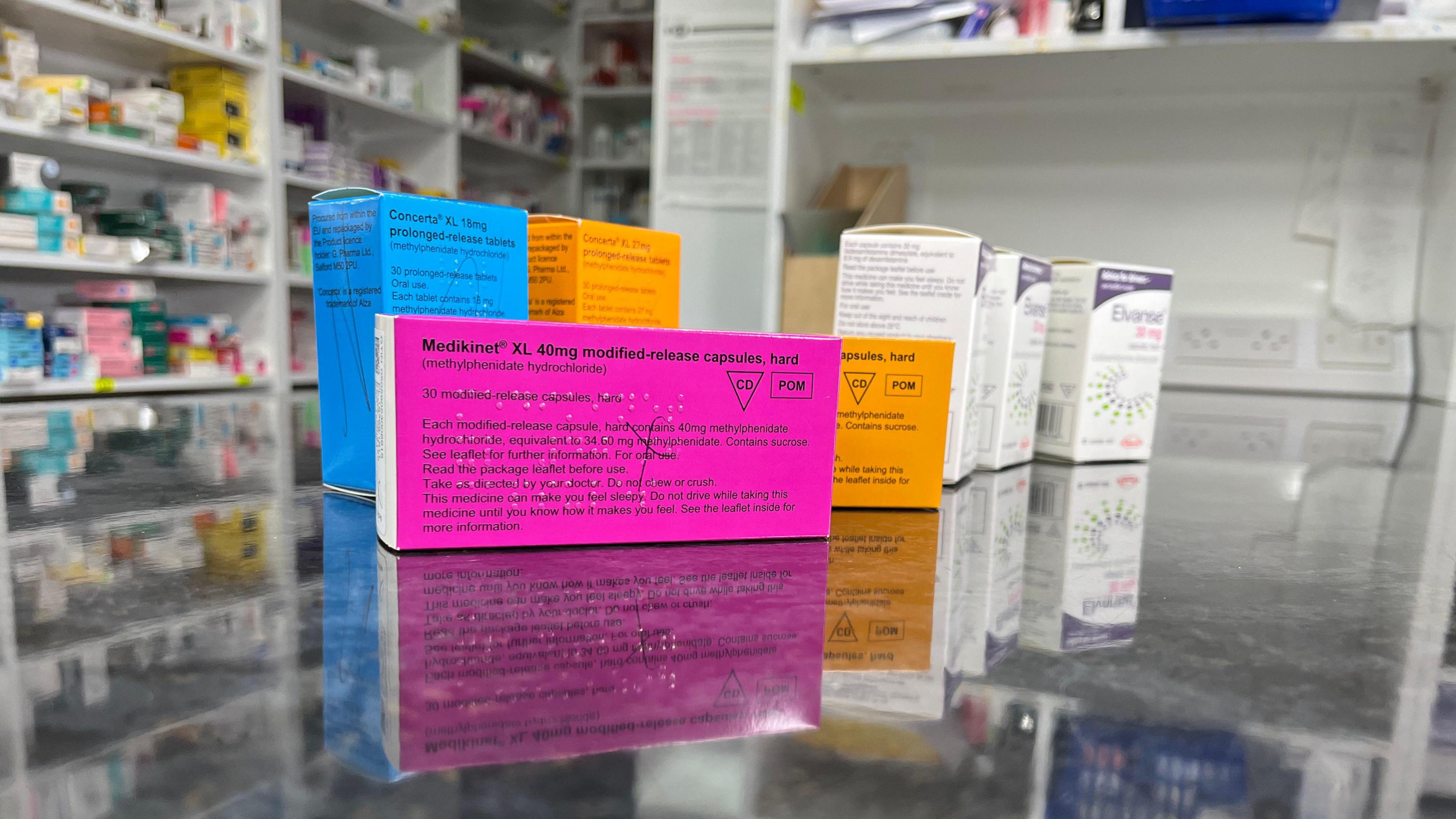 Seven cardboard pill boxes are in the foreground of the photo - one is pink with black writing, two are orange, one is blue and another three are white. They are resting on a reflective pharmacy counter.