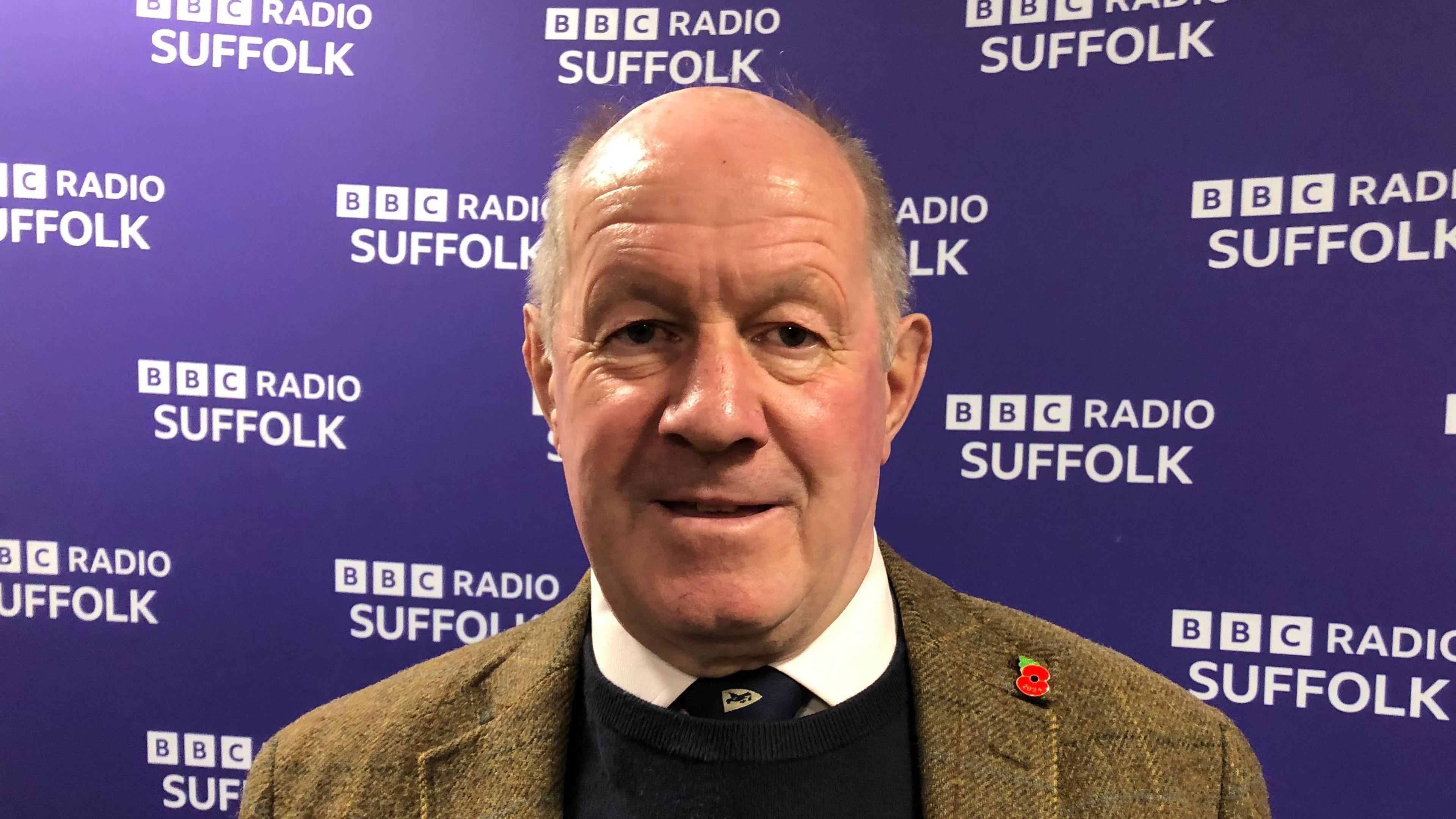 Tim Passmore, wearing a green blazer over a white shirt, black tie and black jumper, standing in front of the BBC Radio Suffolk purple backdrop.