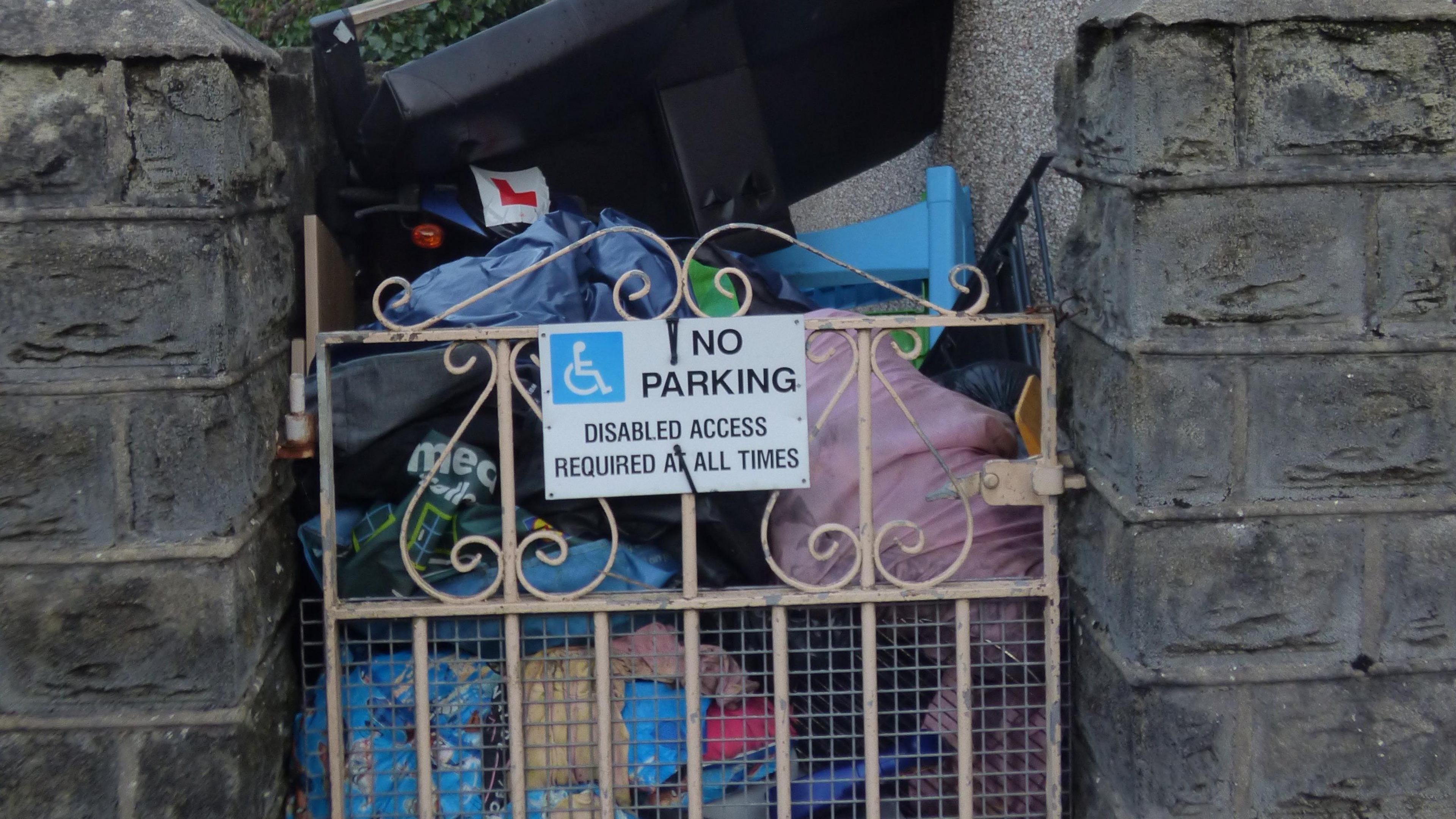 A mound of rubbish stacked behind a small metal gate which bears a sign saying 'NO PARKING: Disabled access required at all times'.
