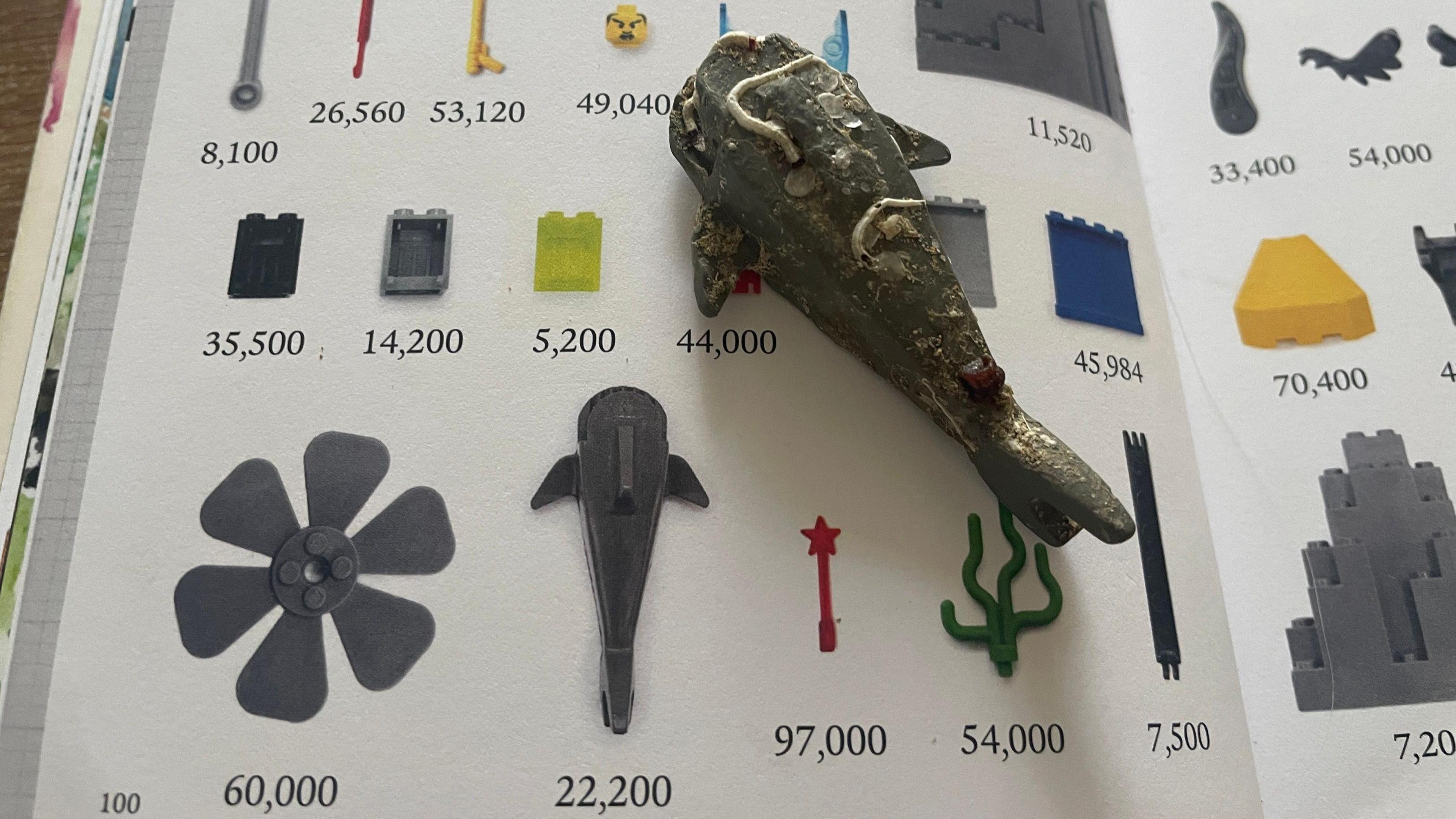 The grey Lego shark lying on a book which has illustrations of some of the lost pieces of Lego, together with numbers showing how many were lost