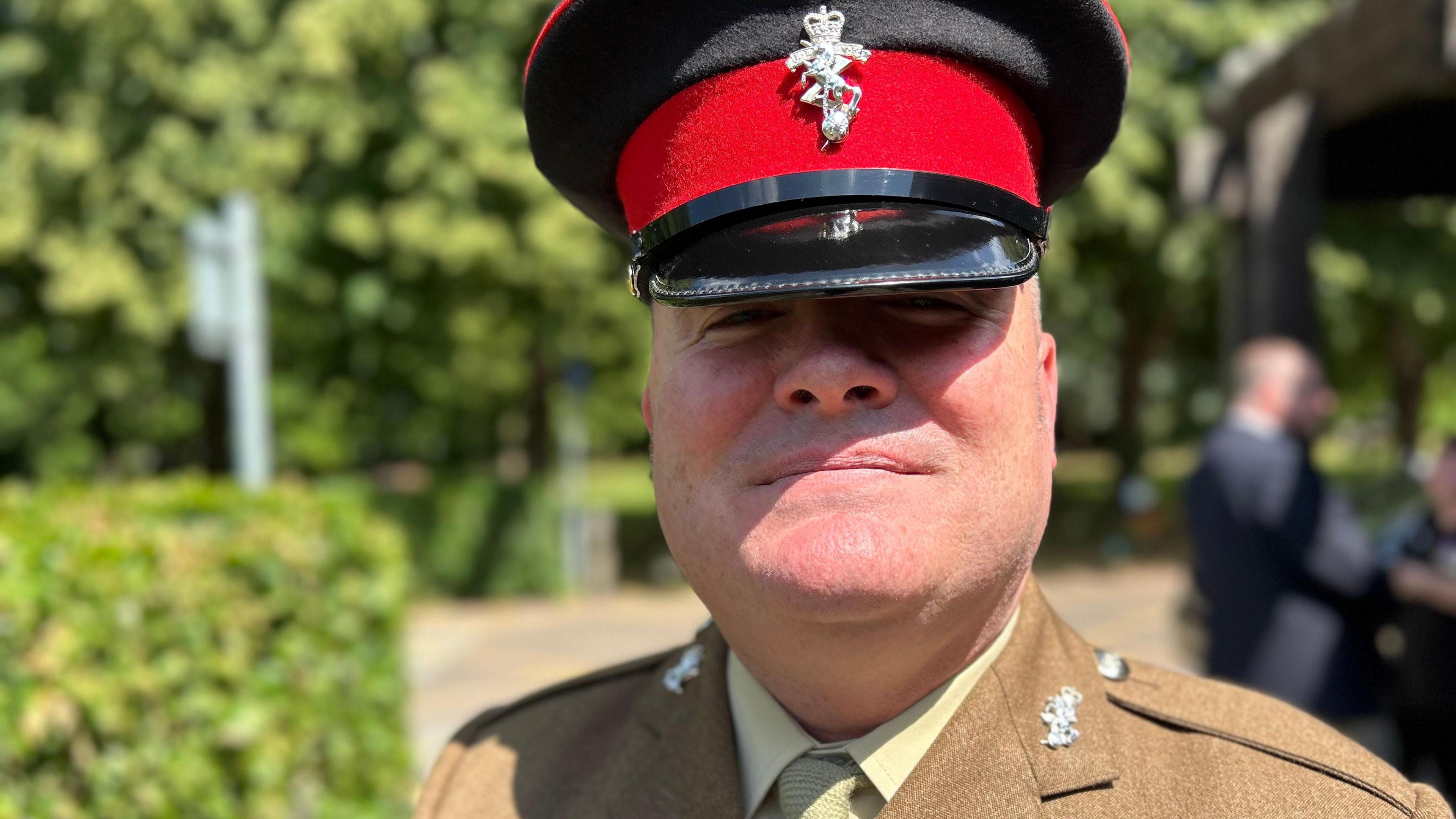 A man wearing an Army uniform with khaki jacket and light-green shirt and tie, along with a black and red cap with a silver badge. He is standing in the grounds of a crematorium.
