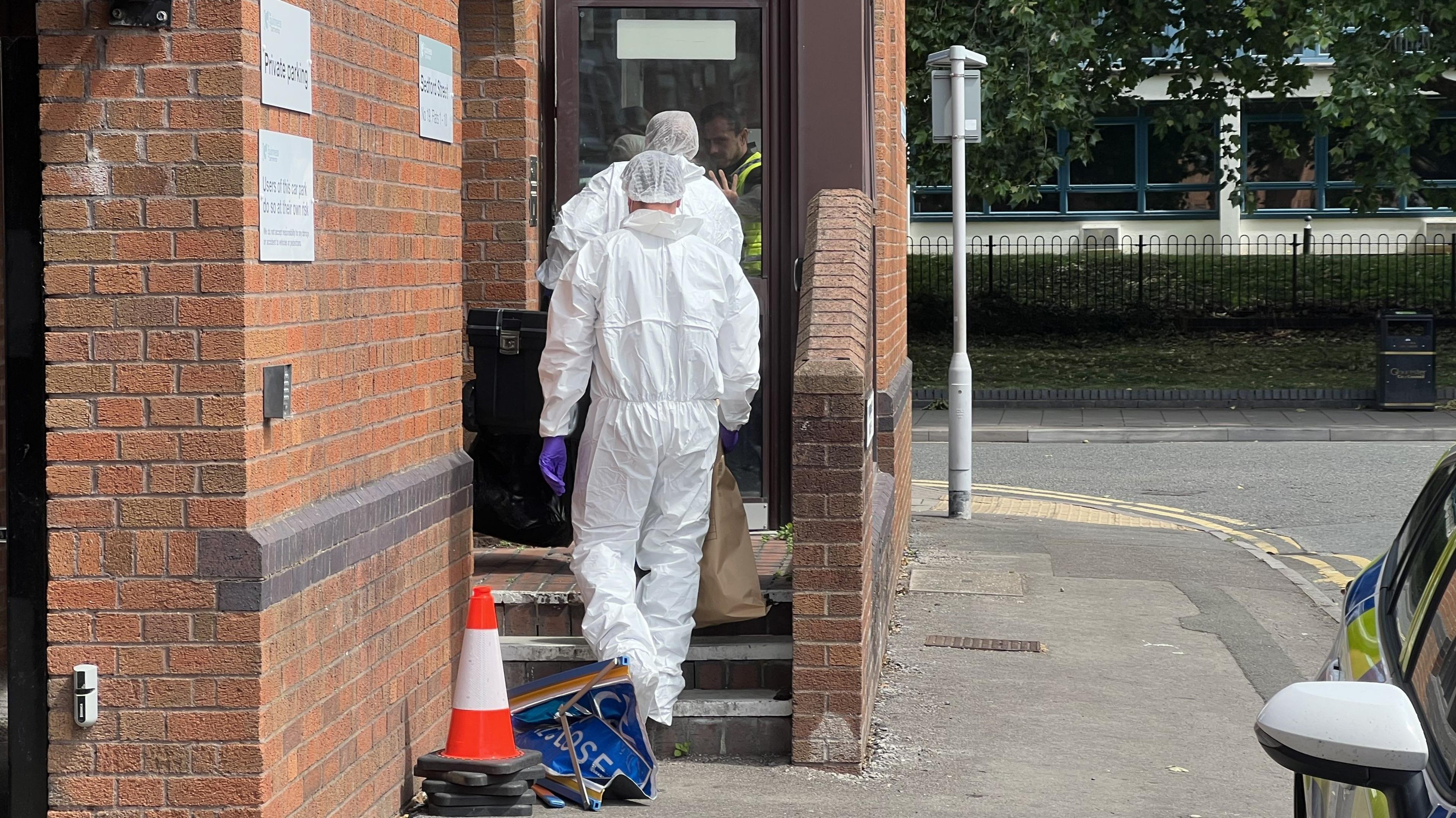 Two people wearing white forensic suits and purple gloves entering a brick building on Bedford Street. One of them is carrying a brown paper bag as he walks up the stairs into the property. A police car can be seen parked to the right.