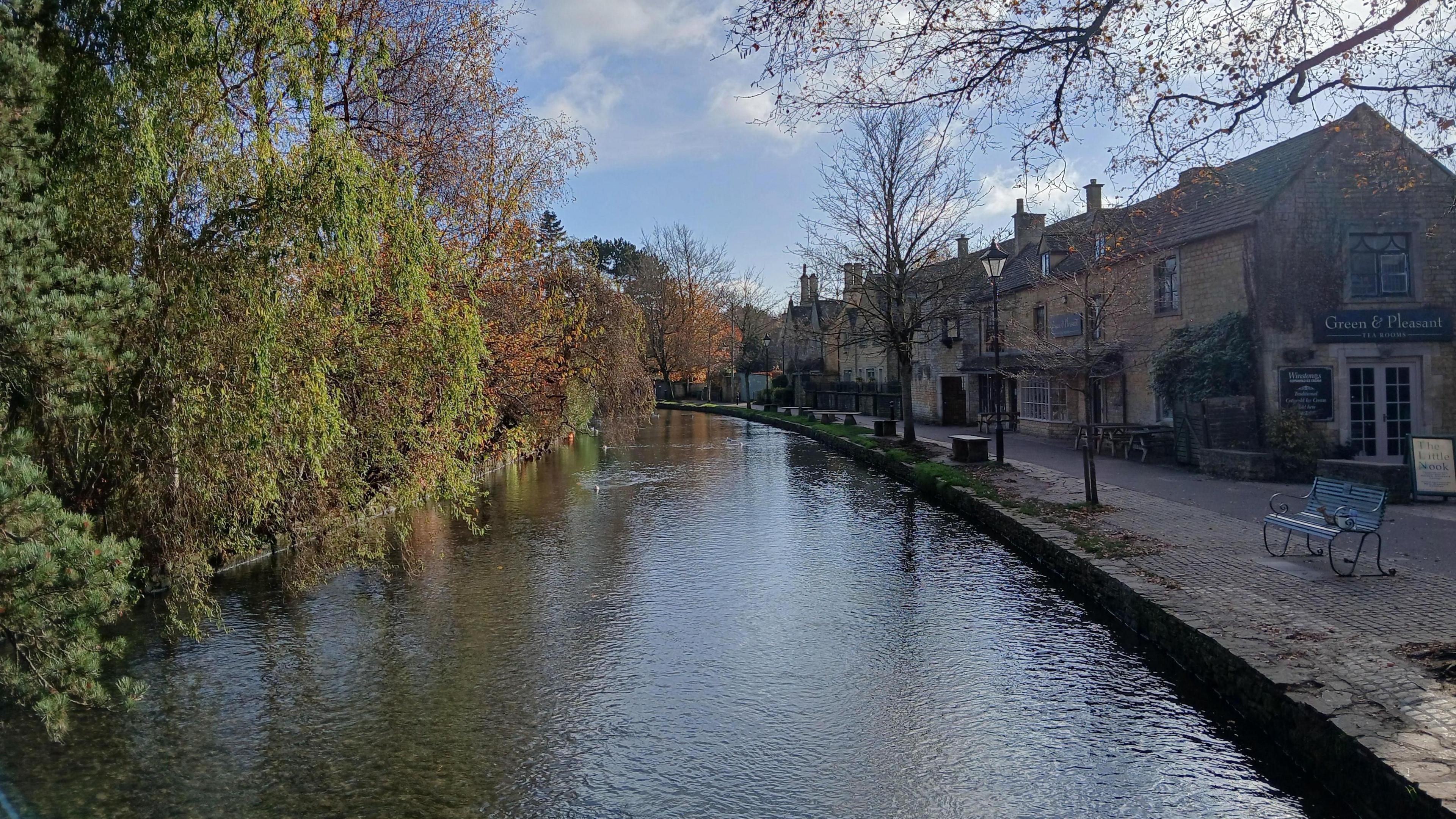 Cotswold brick buildings along the side of the river at Bourton on the Water with green trees on the side. 