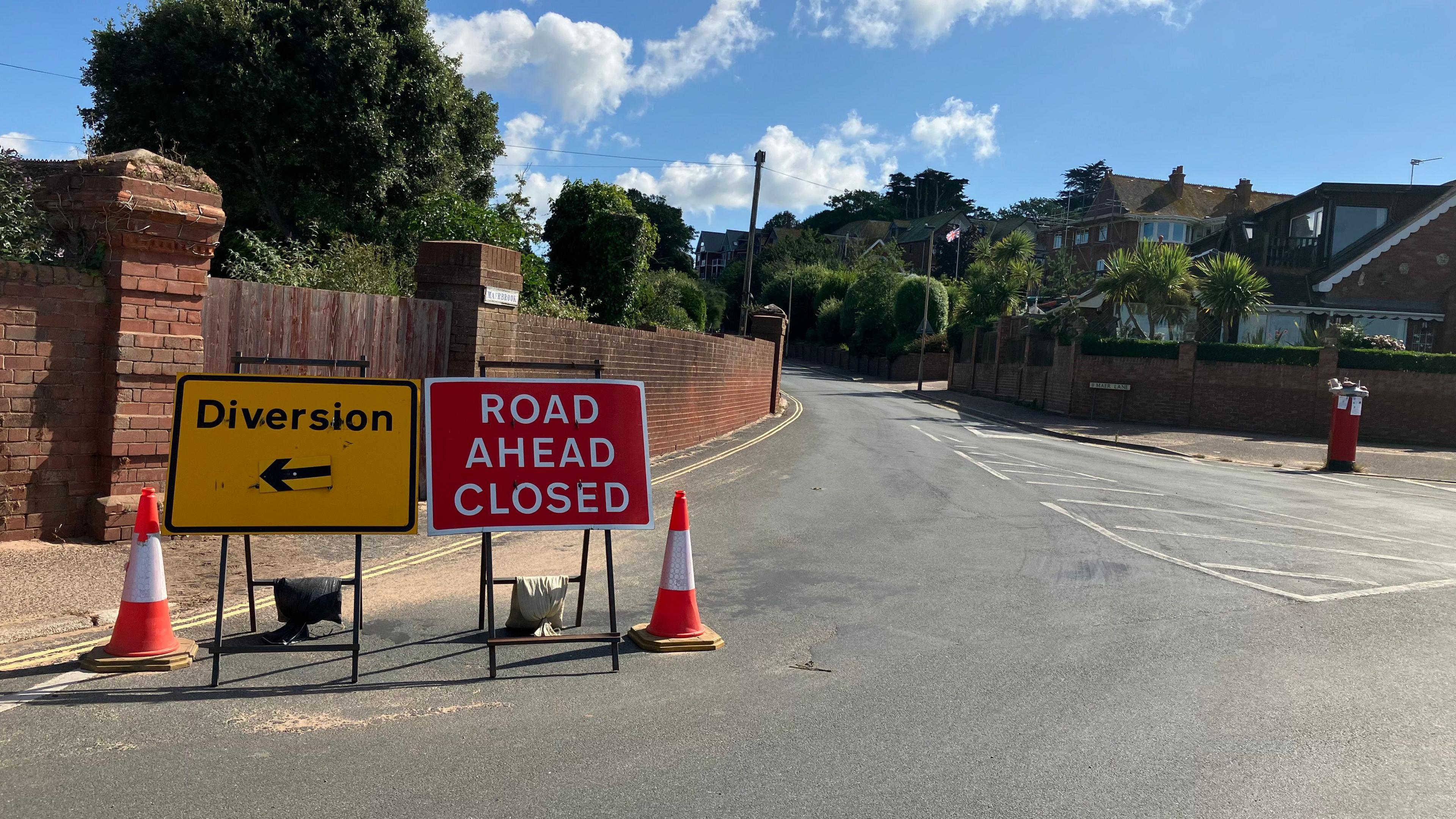 Road closed and diversion signs by the entrance to a road with walls, plants and housing visible either side of the road