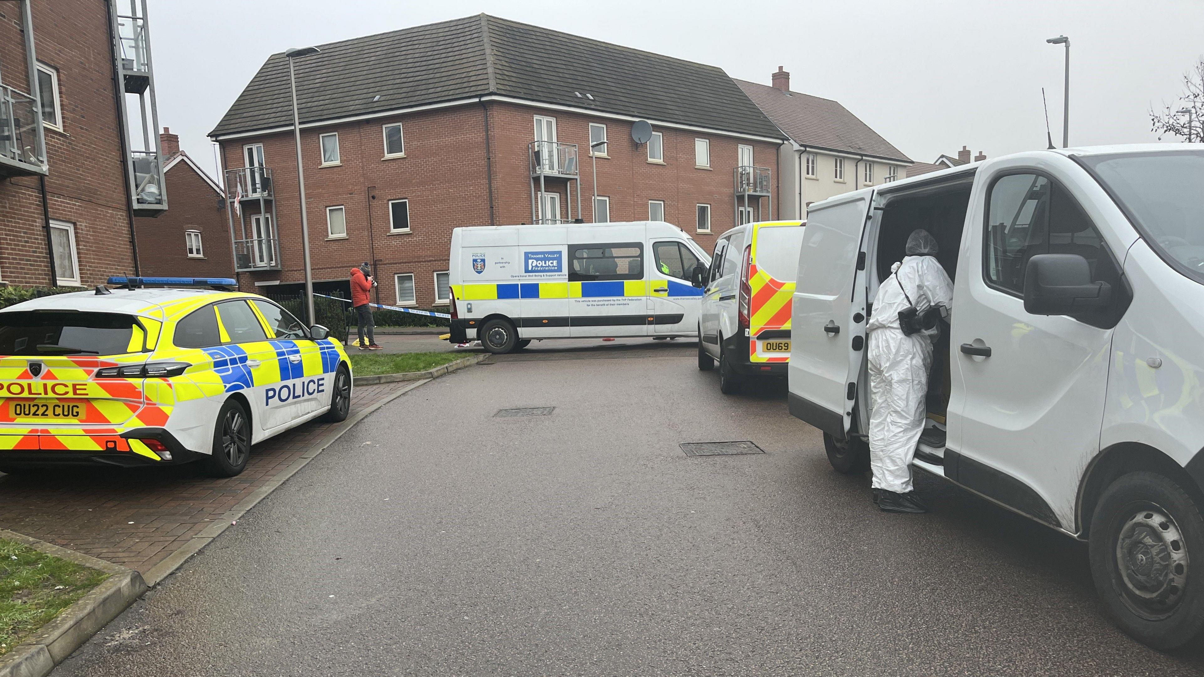 A police car and two police vans along with a white forensic van are parked on a residential street in front of a police cordon. Blocks of flats are pictured in the distance. A forensic officer can be seen looking into the site of the white van.