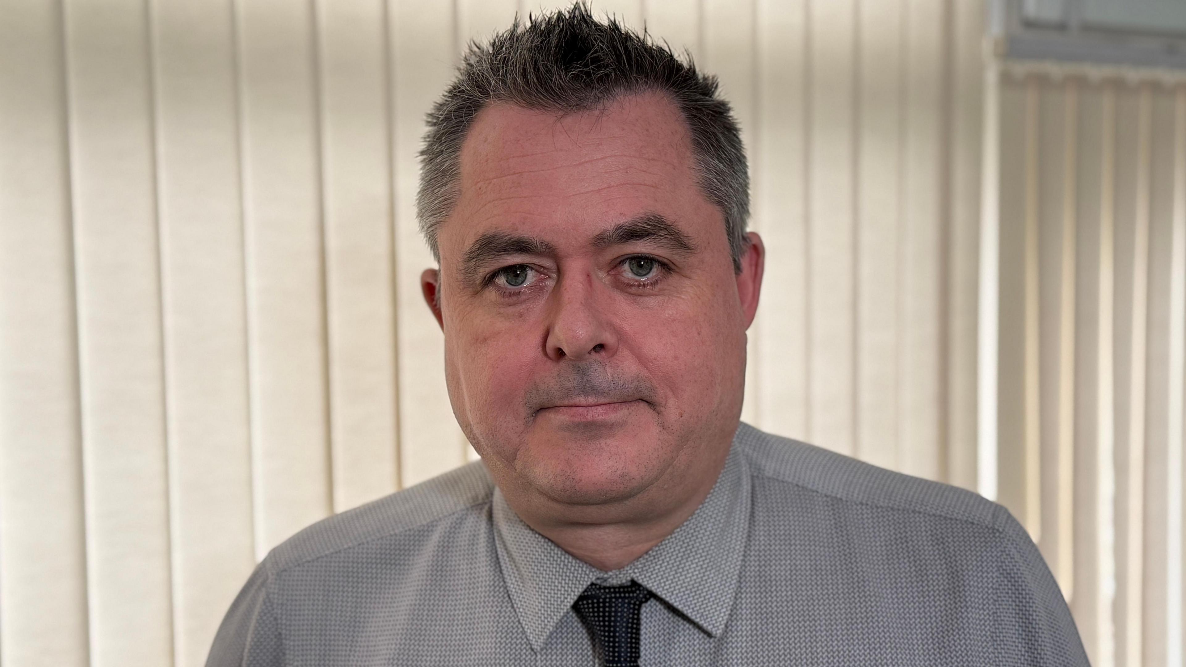 A man with short dark hair that is greying at the side looks straight at the camera. He is wearing a grey shirt and a black tie. The background is made up of cream-coloured blinds.