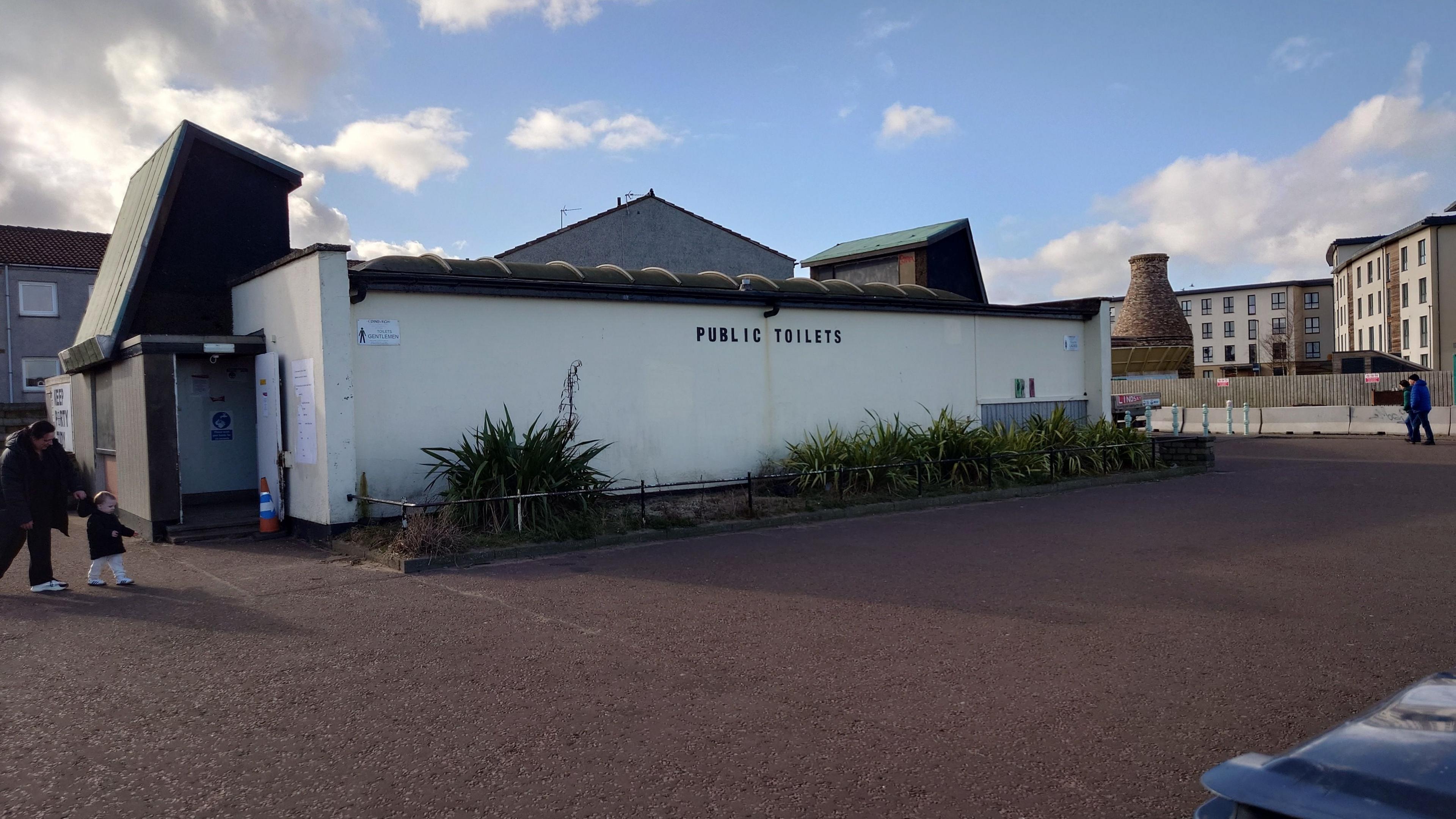 A white public toilet block. There are buildings in the background and a paved area in the foreground.