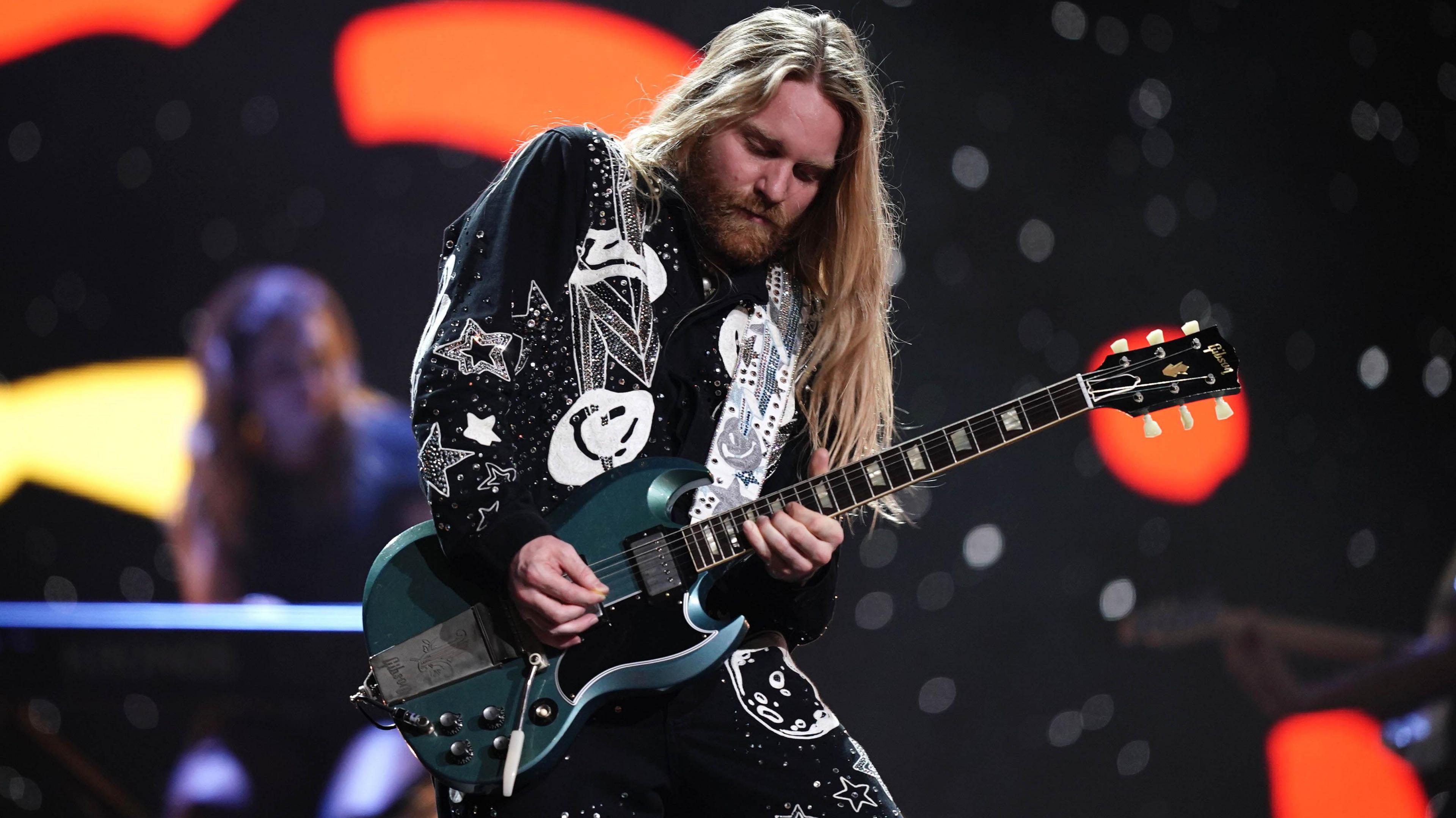 Same Ryder, who has long blonde hair and is wearing a black and white sparkly outfit, playing an electric guitar on stage.