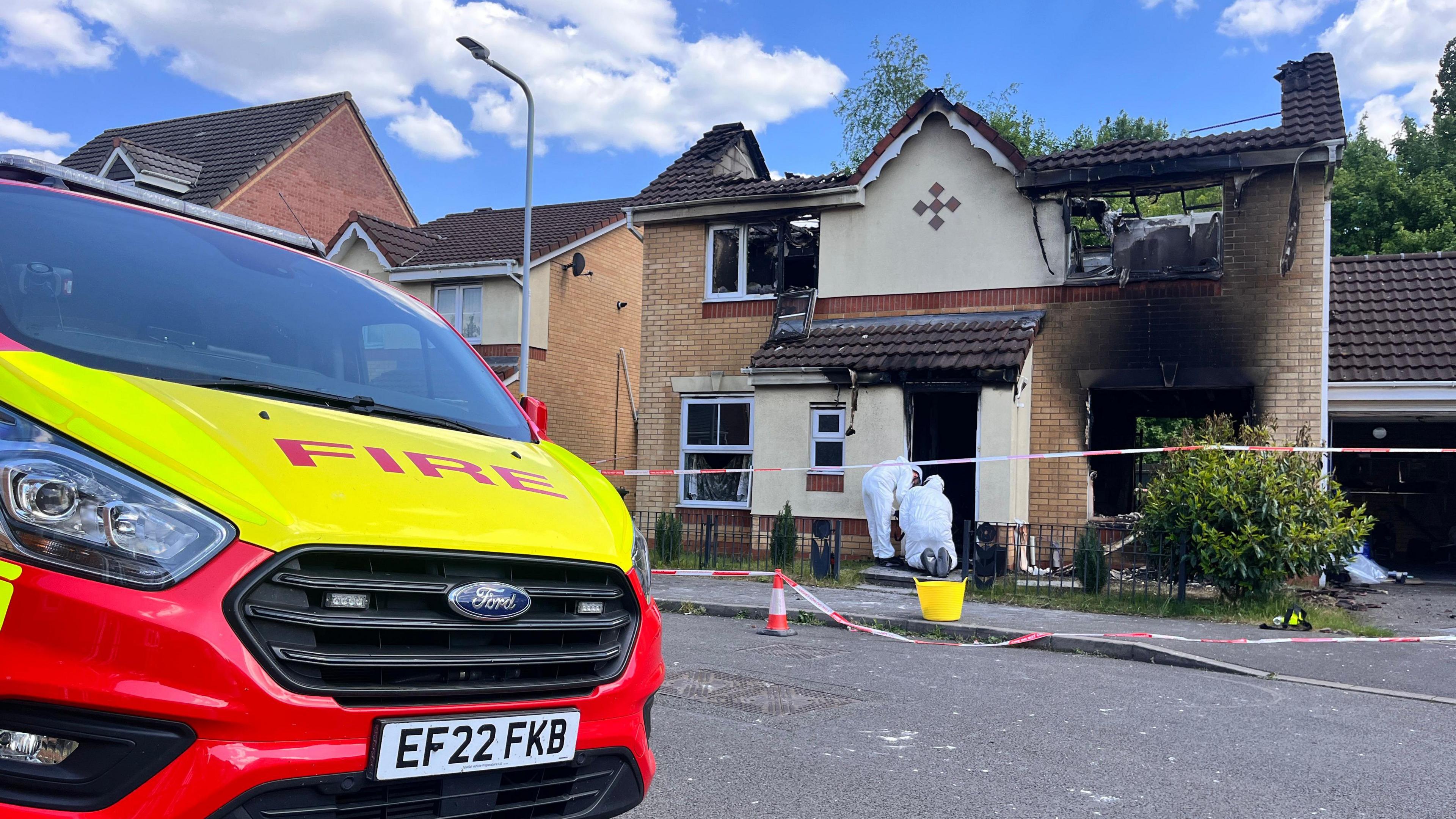 Forensic officers wearing white suits are outside a house that has been destroyed in a fire. It is a burnt-out shell with views through to the back garden. The forensic officers are working where the front door would have been. Red and white fire tape is outside. In the foreground there is a large vehicle with the word "FIRE" in red on a yellow bonnet.