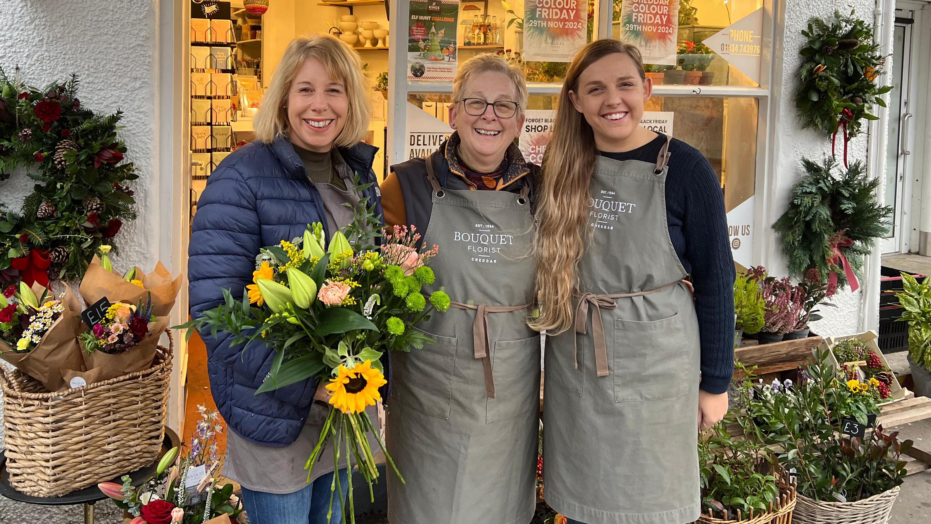 Claire Wilcox stands on the right, next to two of her colleagues who run a flower shop in Cheddar. Claire is holding a bouquet. Behind them is the entrance to their shop, with flowers displayed outside.