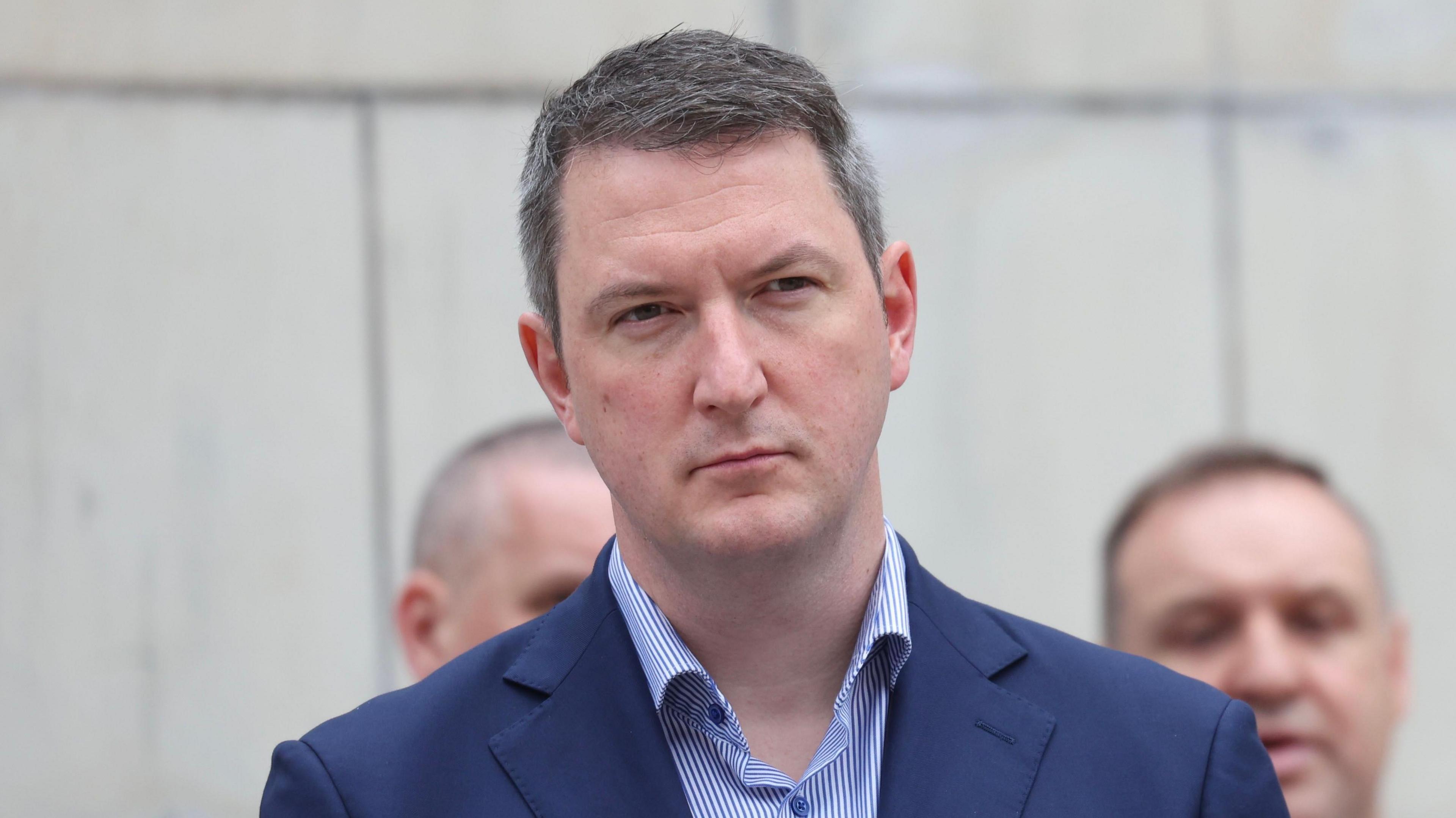 A still of John Finucane standing outside a white-stone building. He has short greying hair and is wearing a blue suit jacket as well as a blue and white striped shirt. The background is blurred.