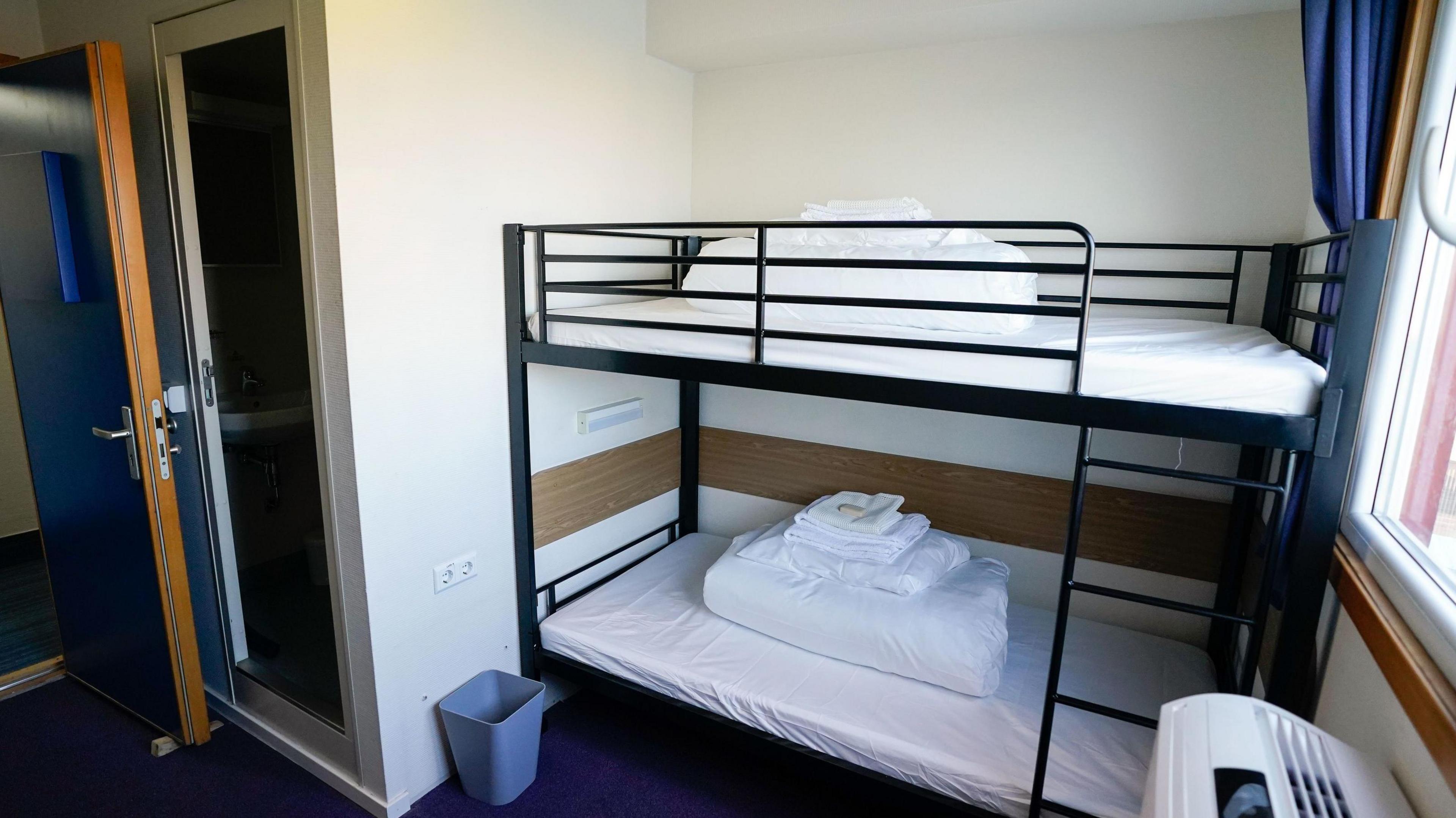 A black bunk bed with white sheets, in a small room with white walls and a blue floor.