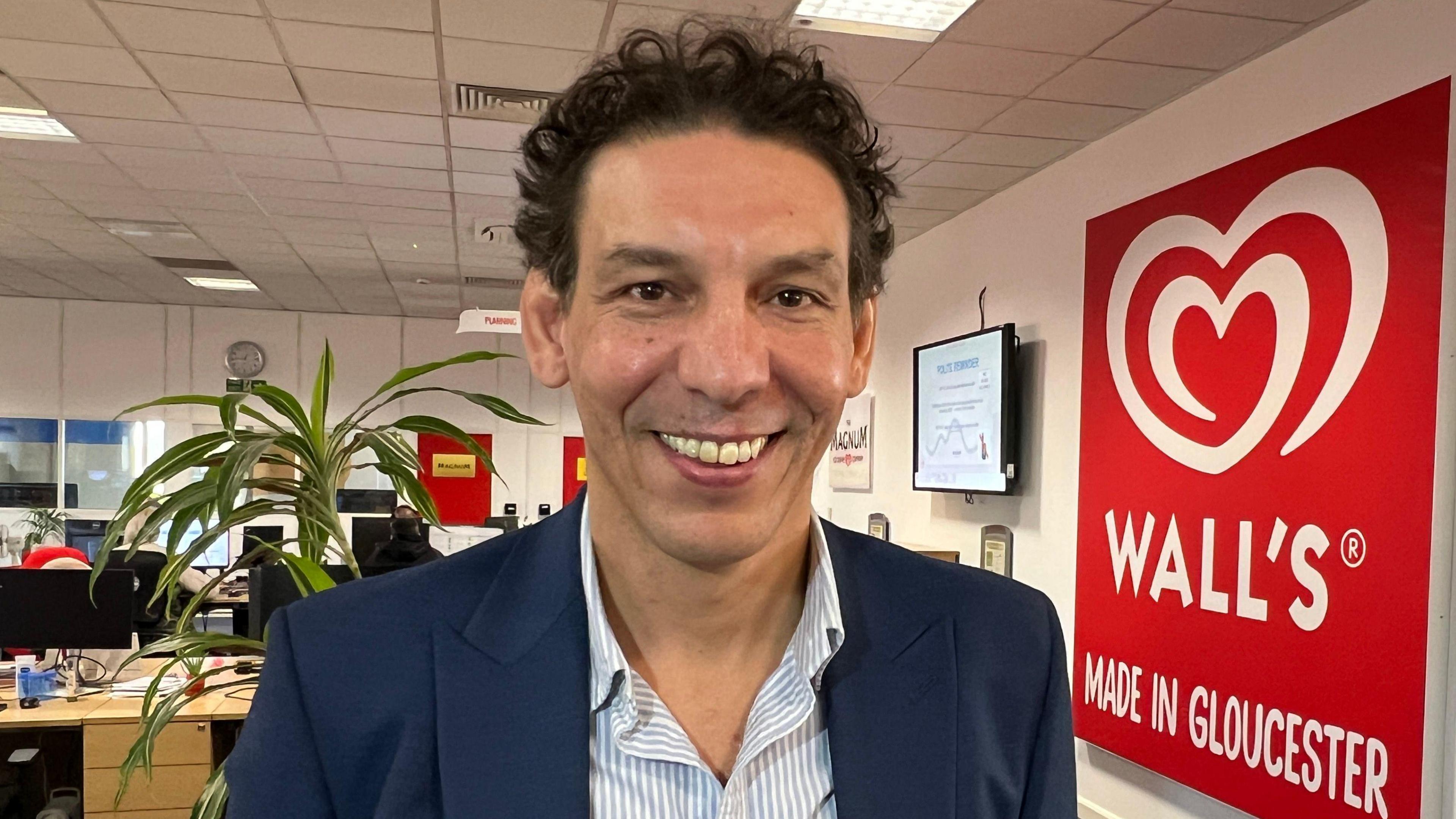 A man is standing in an office wearing a blue suit. He is smiling at the camera. Behind him is a red sign which has white writing which says 'Wall's Made in Gloucester'. 