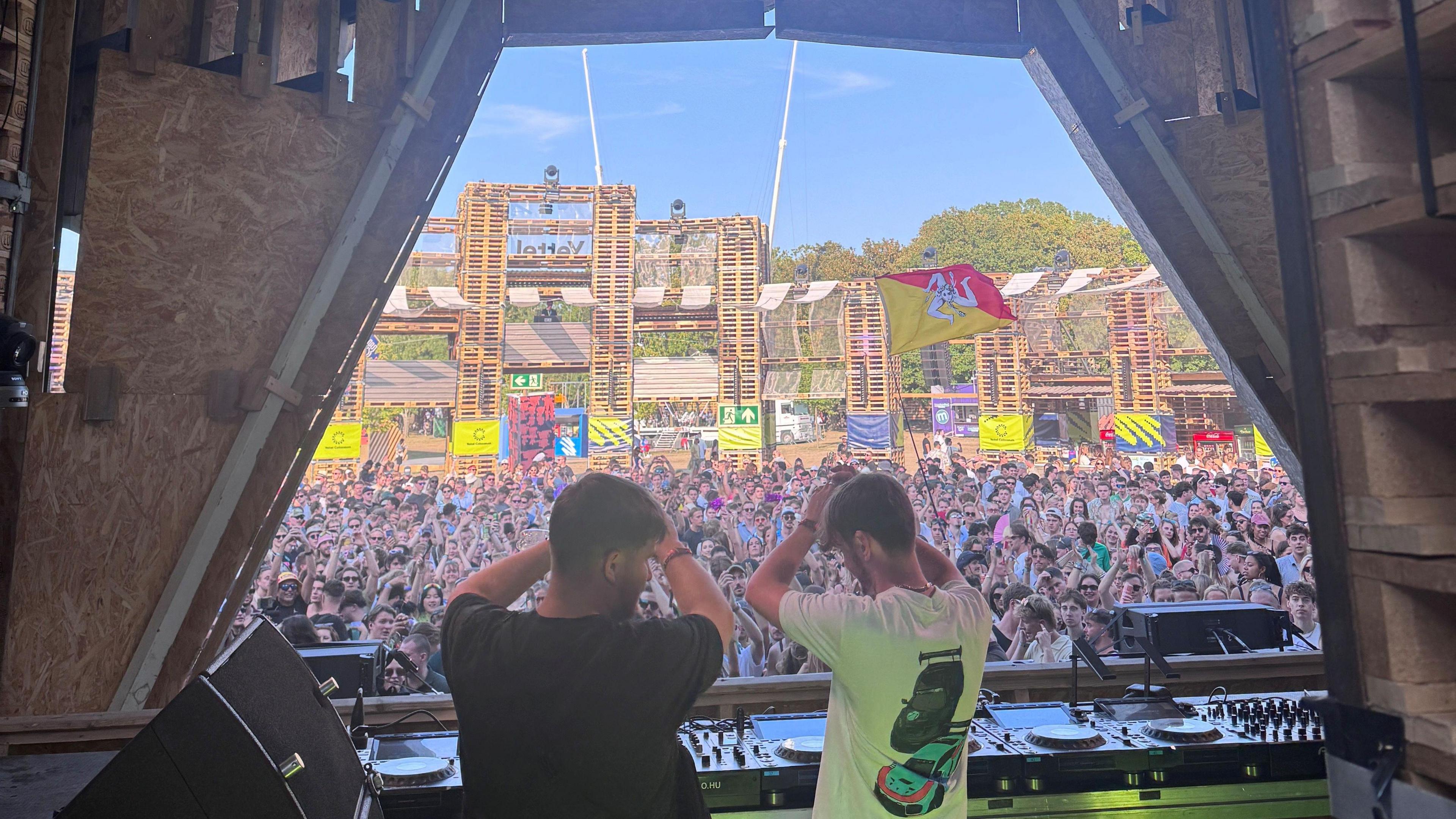 Two men hold their hands up as they stand behind decks at a music show. A crowd can be seen in the background cheering them on.