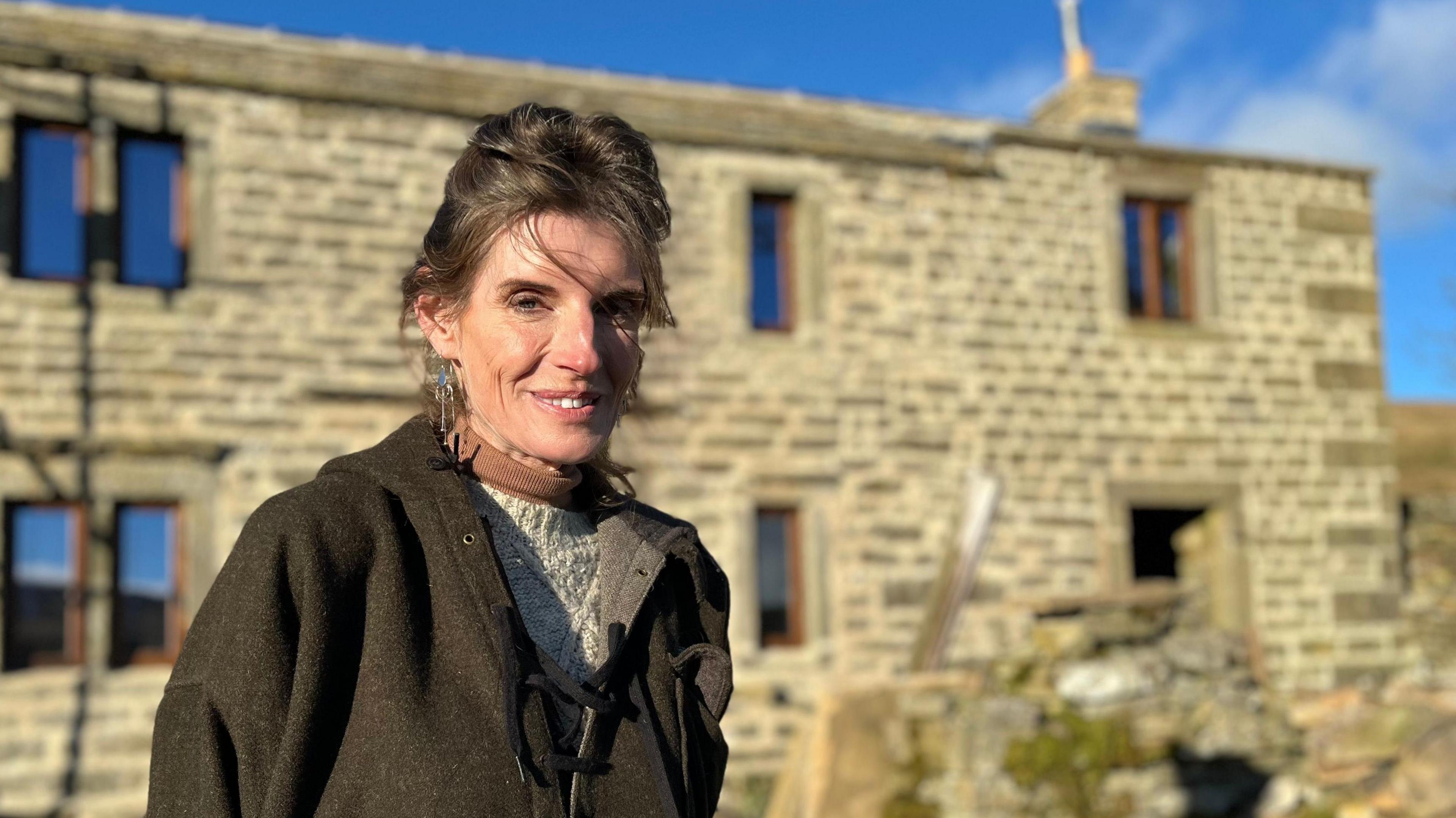 Amanda Owen has dark hair tied back and wears a green tweed jacket as she stands in front of a stone farmhouse.