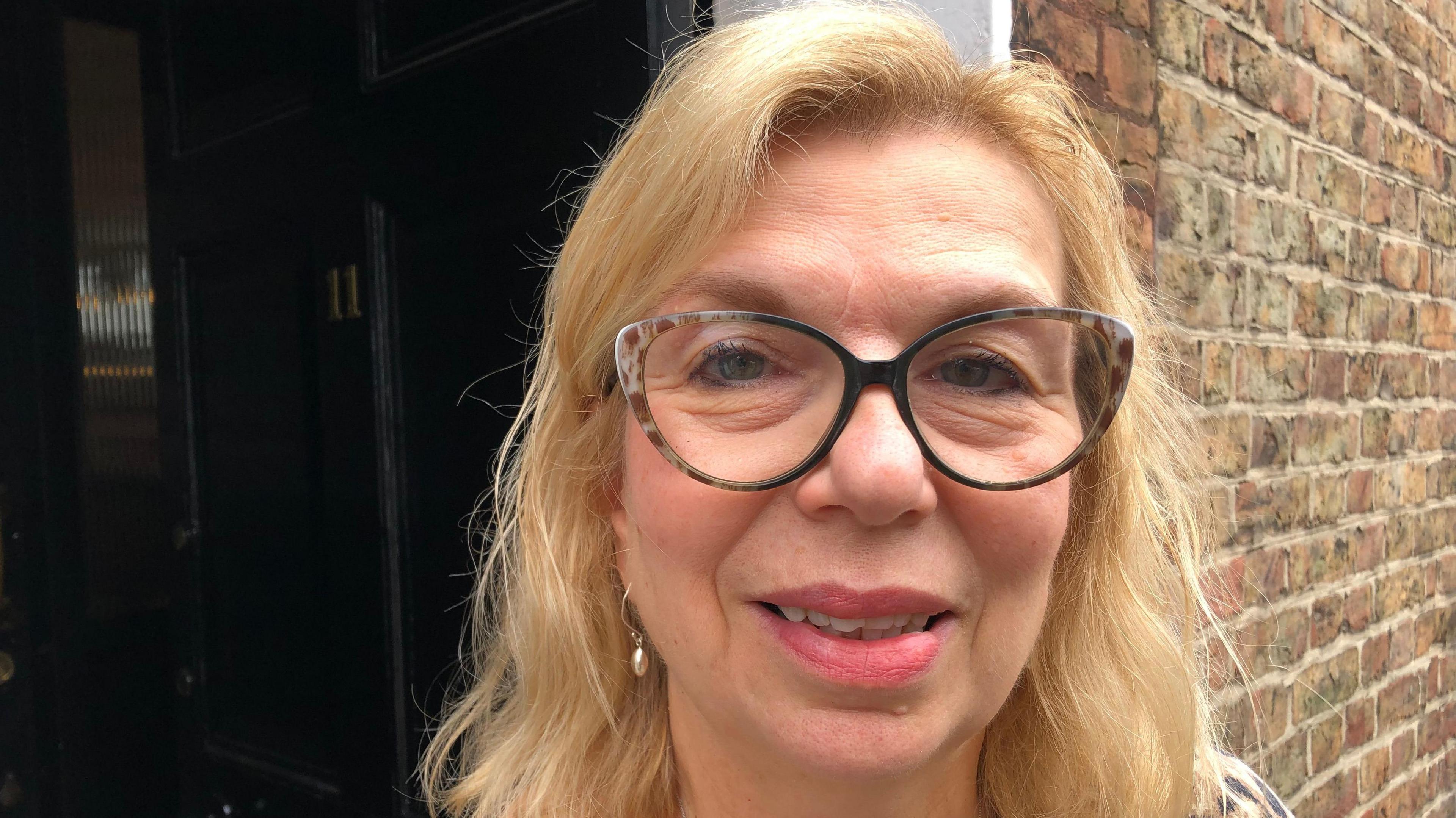 Close-up image of hairdresser Zoe Kubat next to large black door. She has blond hair and is wearing large glasses.