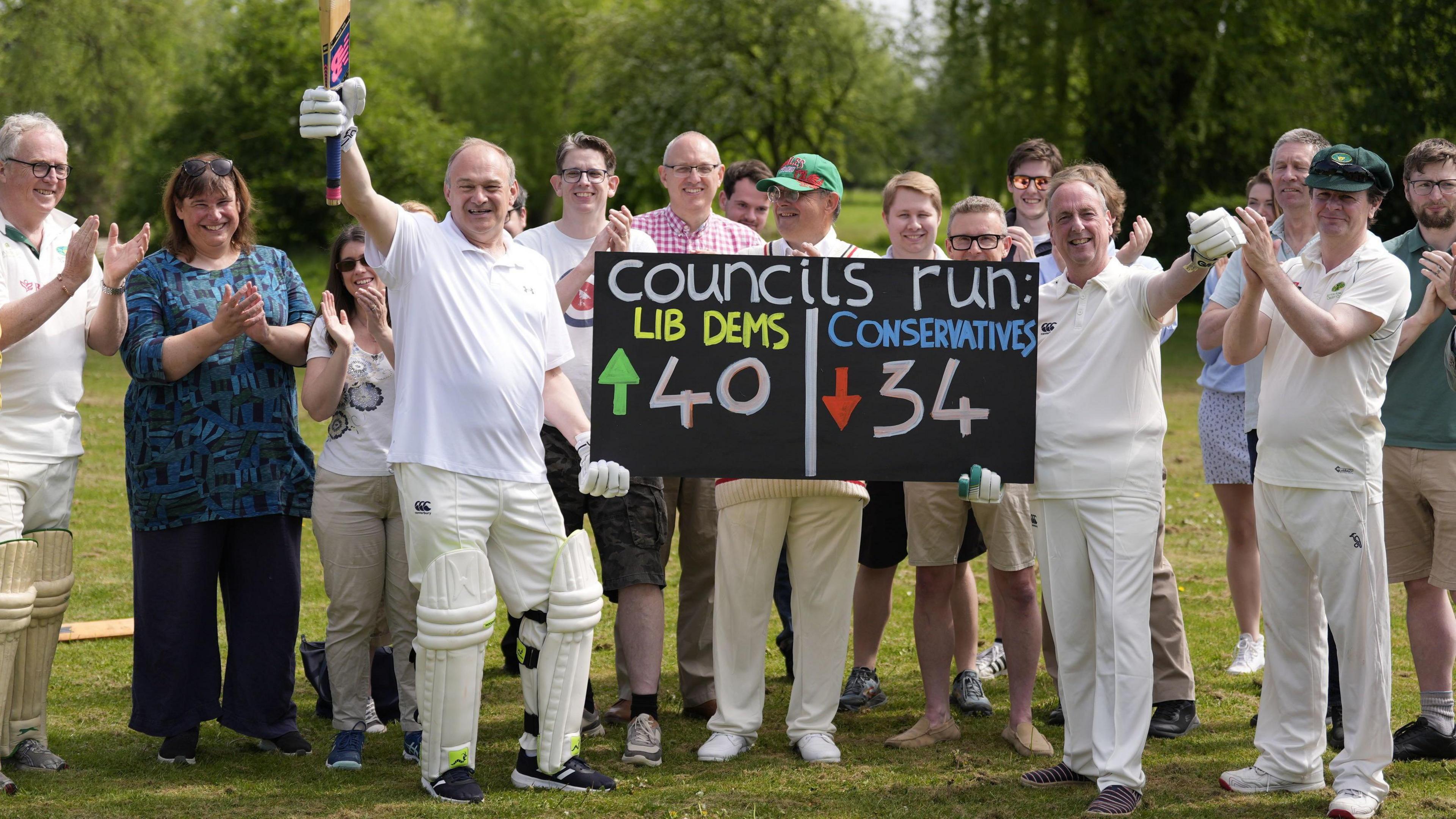 Sir Ed Davey pictured with a group of councillors who are wearing cricket gear. Sir Ed is holding a cricket bat up in the air, while holding a score board in the other which shows the Lib Dem's have gained 40 seats and the Conservatives have lost 34.