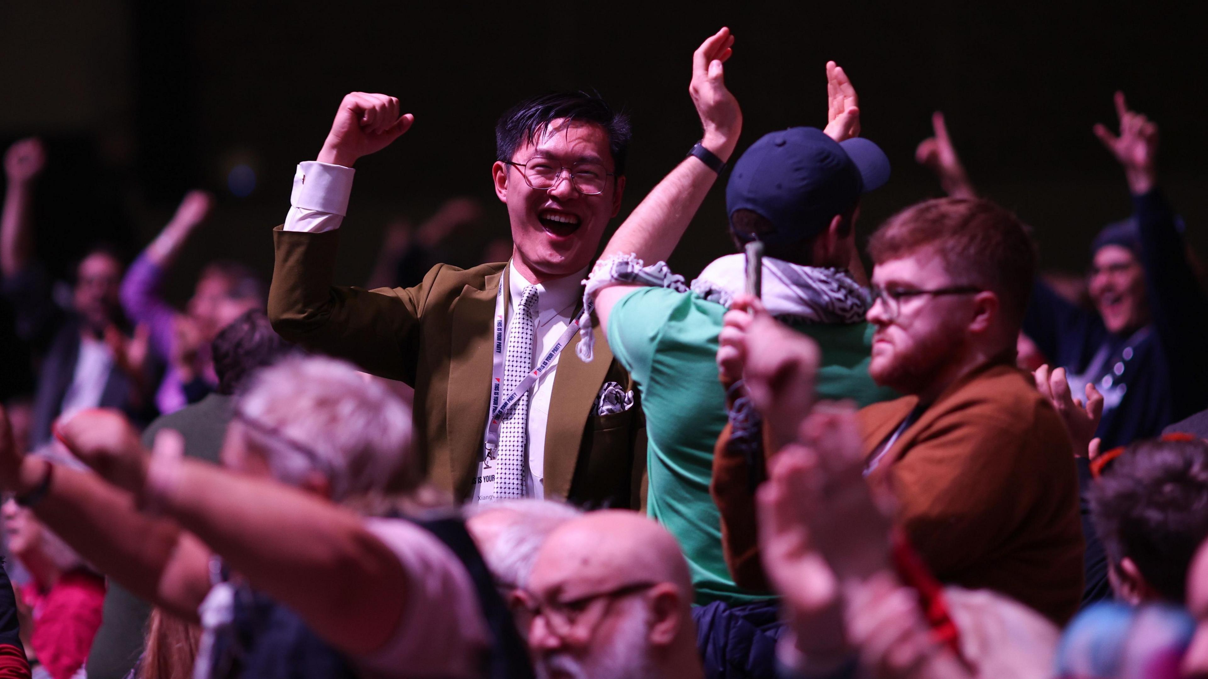 Large crowd of people indoors with raised arms and clenched fists, appearing to celebrate or cheer. Individuals are standing close together, some wearing jackets and scarves, and the background is dimly lit, suggesting a lively event or conference setting.”