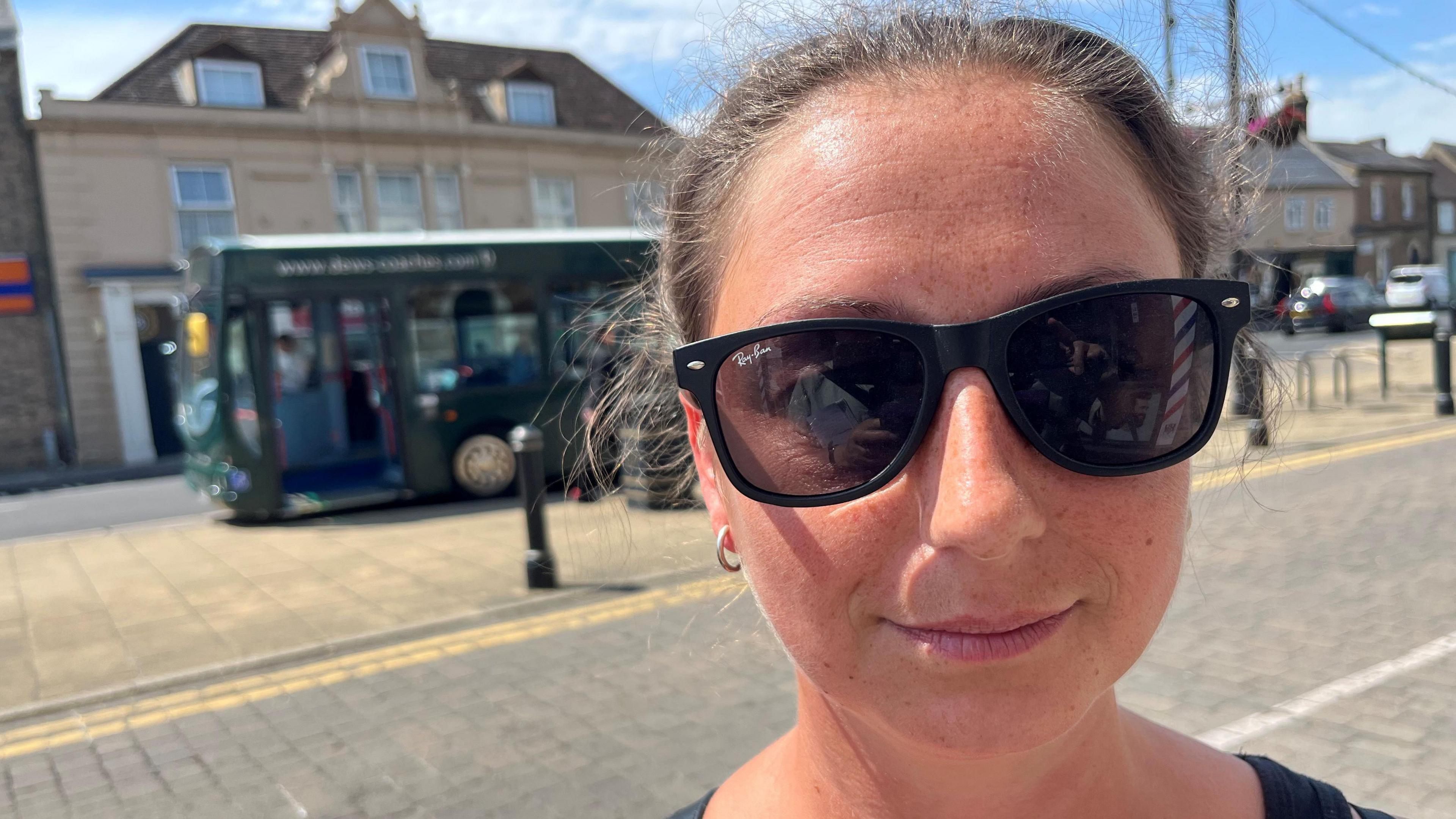 Ashley Whelton, 34, has brown hair pulled back tightly over her head, she is wearing dark glasses in strong sunshine on a street with a green coach at the bus stop behind her, shops and cars can also be seen in the background.