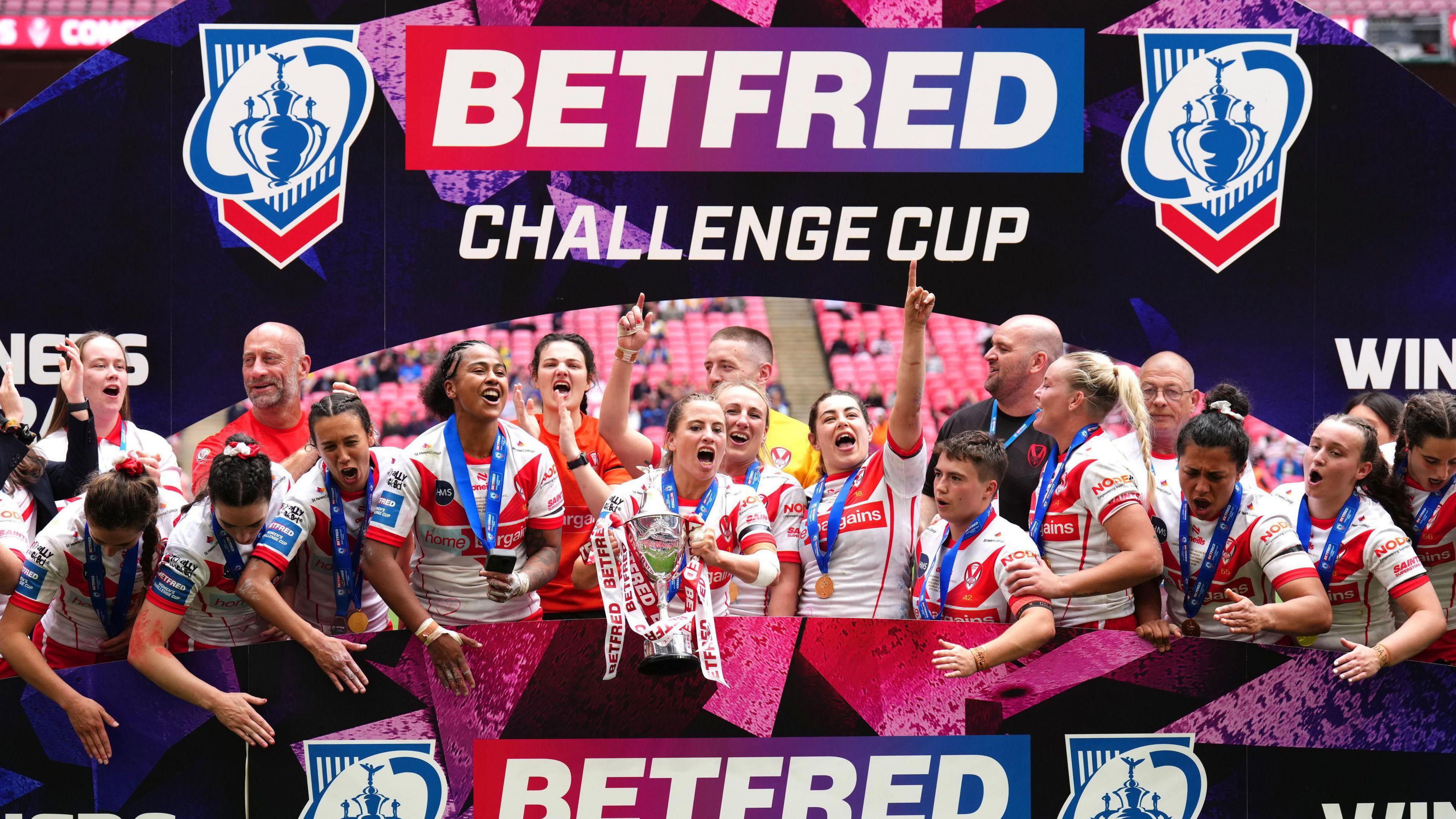 The St Helens players, in their white and red kits and each wearing a blue-ribboned winners medal, lift the trophy in front of blue and red advertising hoardings which read 'BetFred Challenge Cup', after victory in the women's final at Wembley Stadium