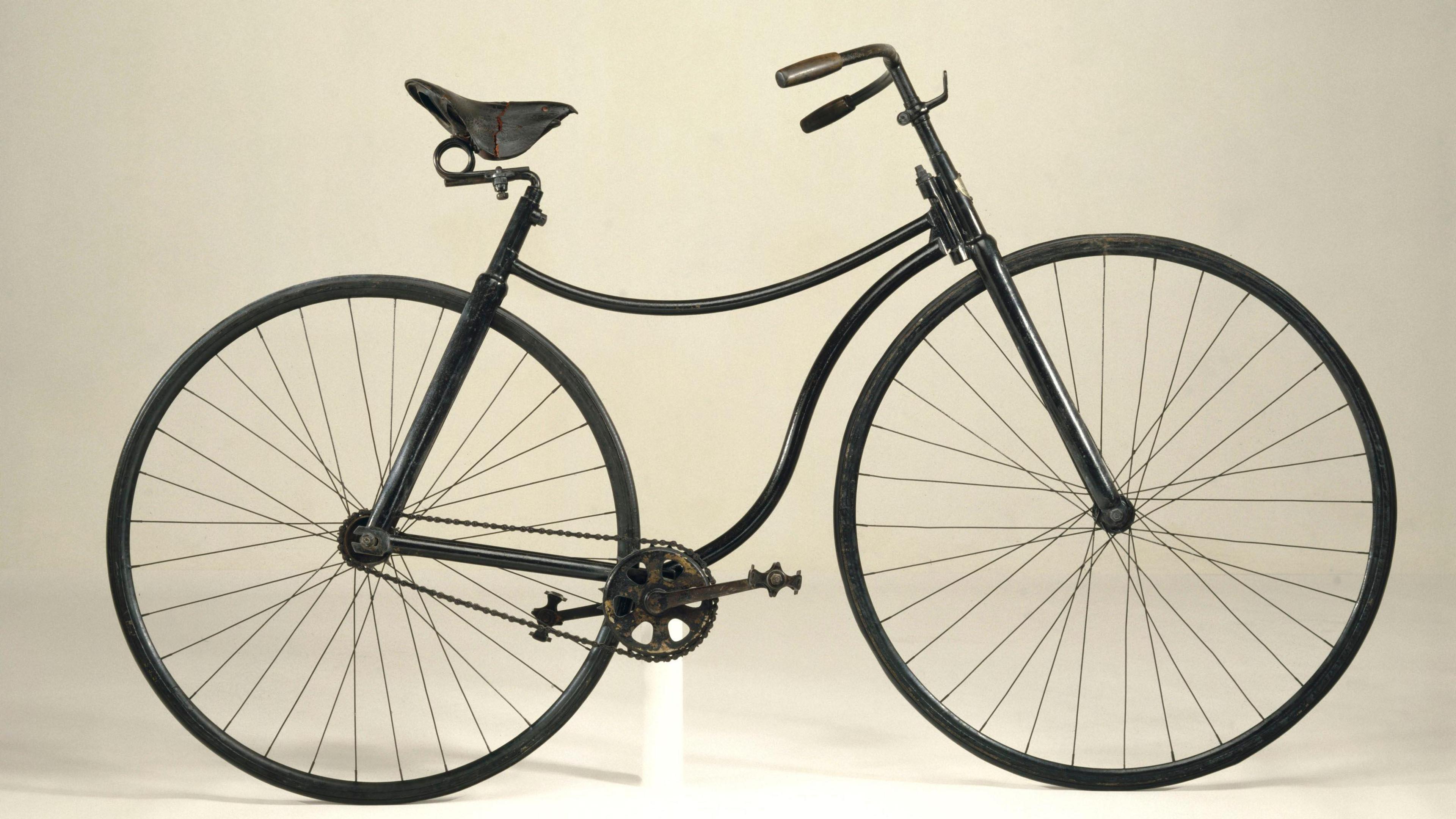 The Rover safety bicycle is an old fashioned black bicycle. We see two wheels with spokes and a gear system between them with pedals.