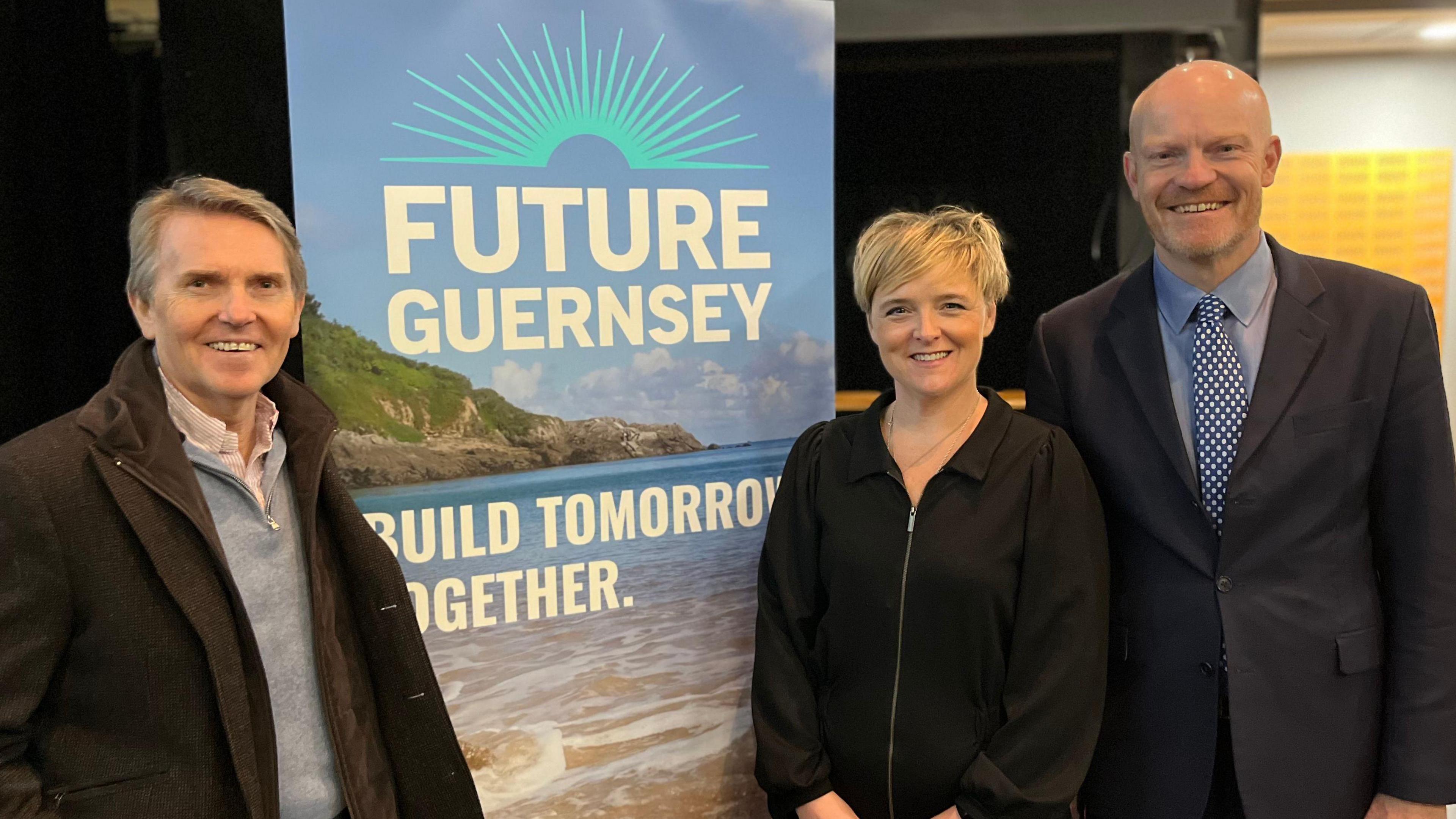 From left to right, a man with grey hair wearing a brown jacket and a grey fleece over a salmon pink shirt smiling towards the camera. On his right is a banner which says Future Guernsey, Build Together Tomorrow. On its right a short blonde lady wearing a black jacket smiling at the camera. On her right is a tall bald man with a ginger beard, wearing a black suit and a blue shirt and blue tie. 