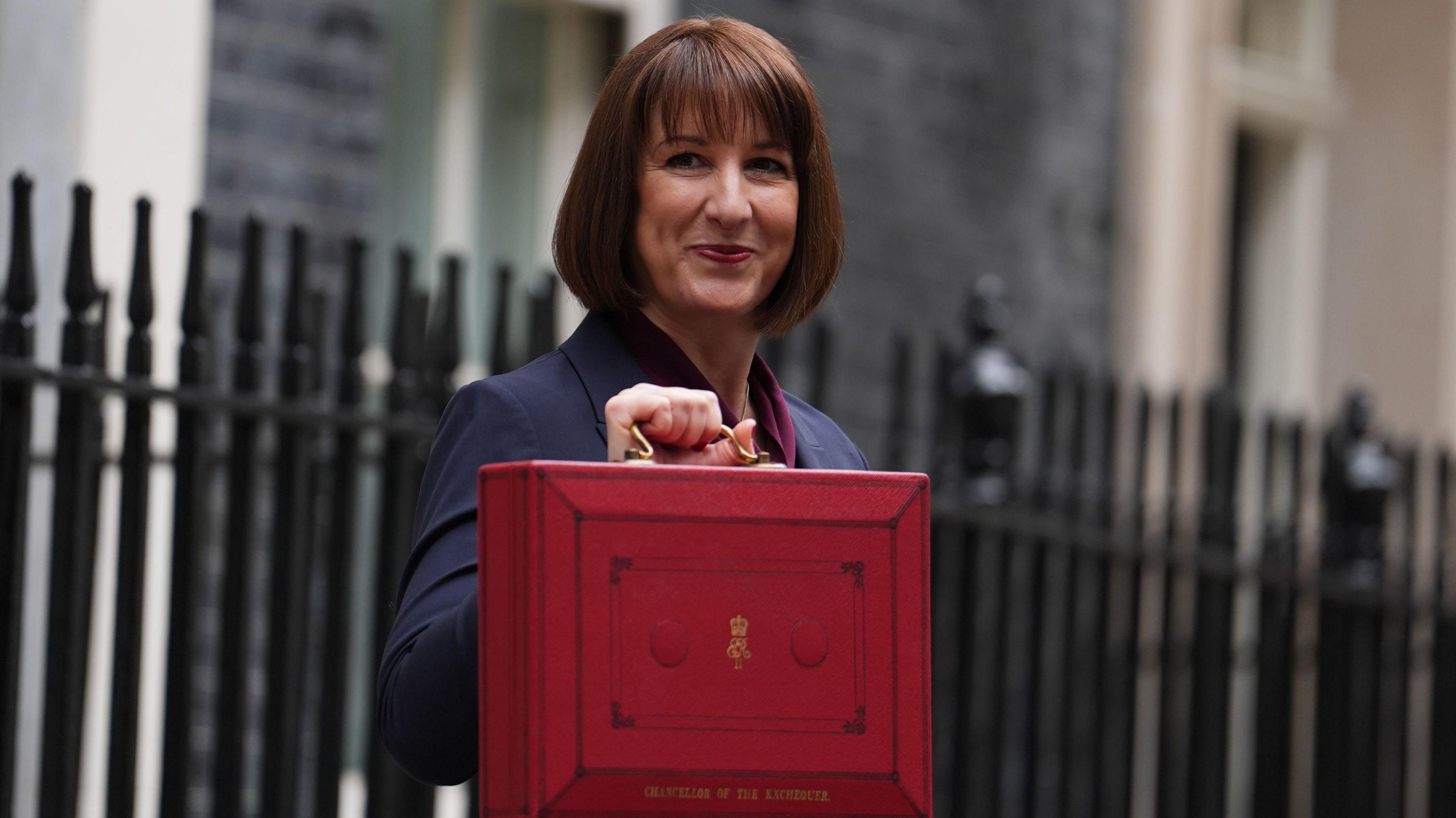 Chancellor Rachel Reeves holds up a red suitcase on Budget Day as she stands on Downing Street. She is slightly smiling as she looks to her right side.