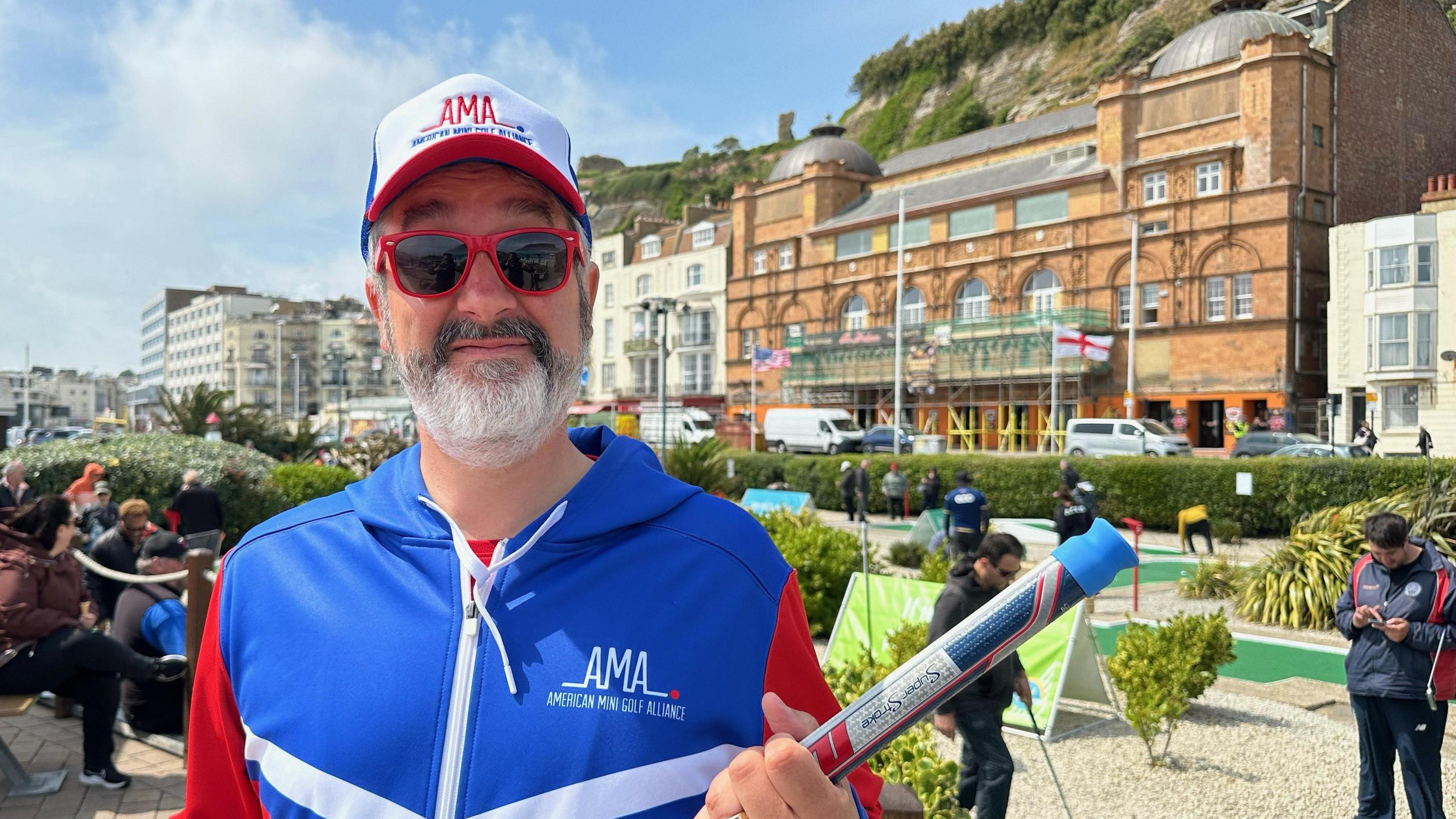 A man dressed in matching red, white and blue zip-up jumper and cap, holding a golf club. He is stood at a crazy gold course in Hastings and is also wearing sunglesses.