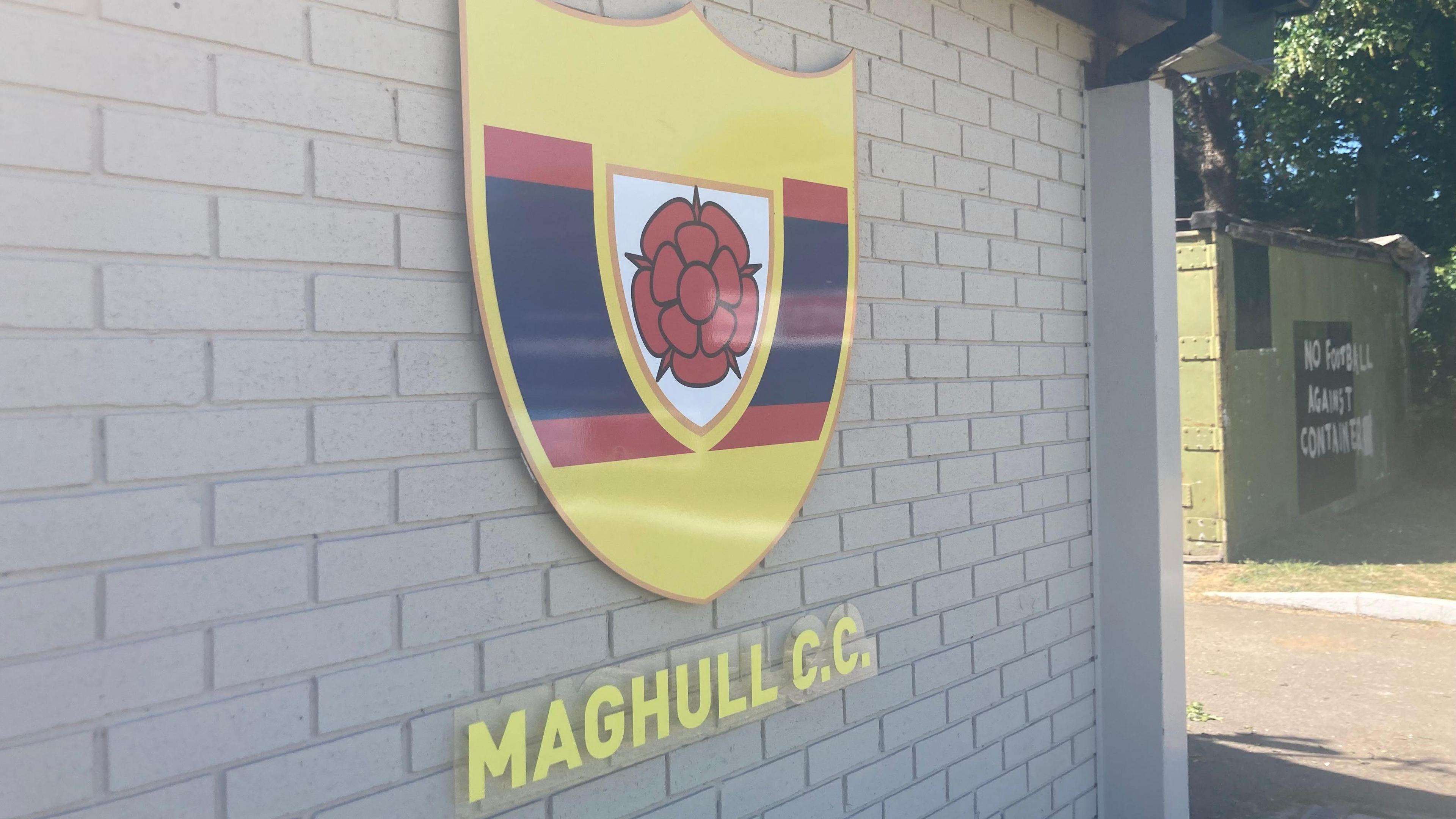 The yellow, black and red logo for Maghull Cricket Club set against a brick wall on a sunny day.