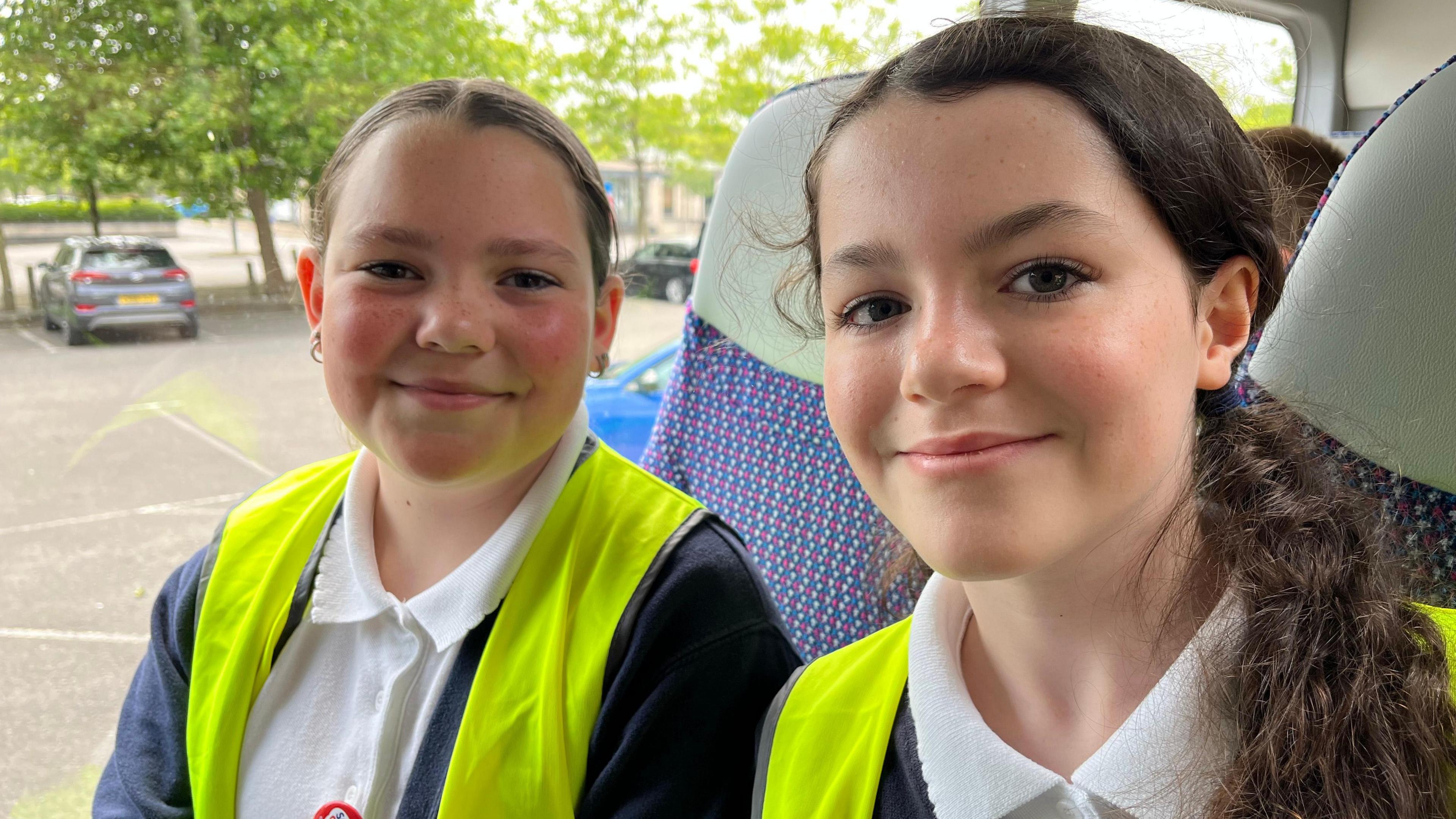 Image of two schoolgirls seated inside a bus. They are wearing high- viz vests over their school uniforms