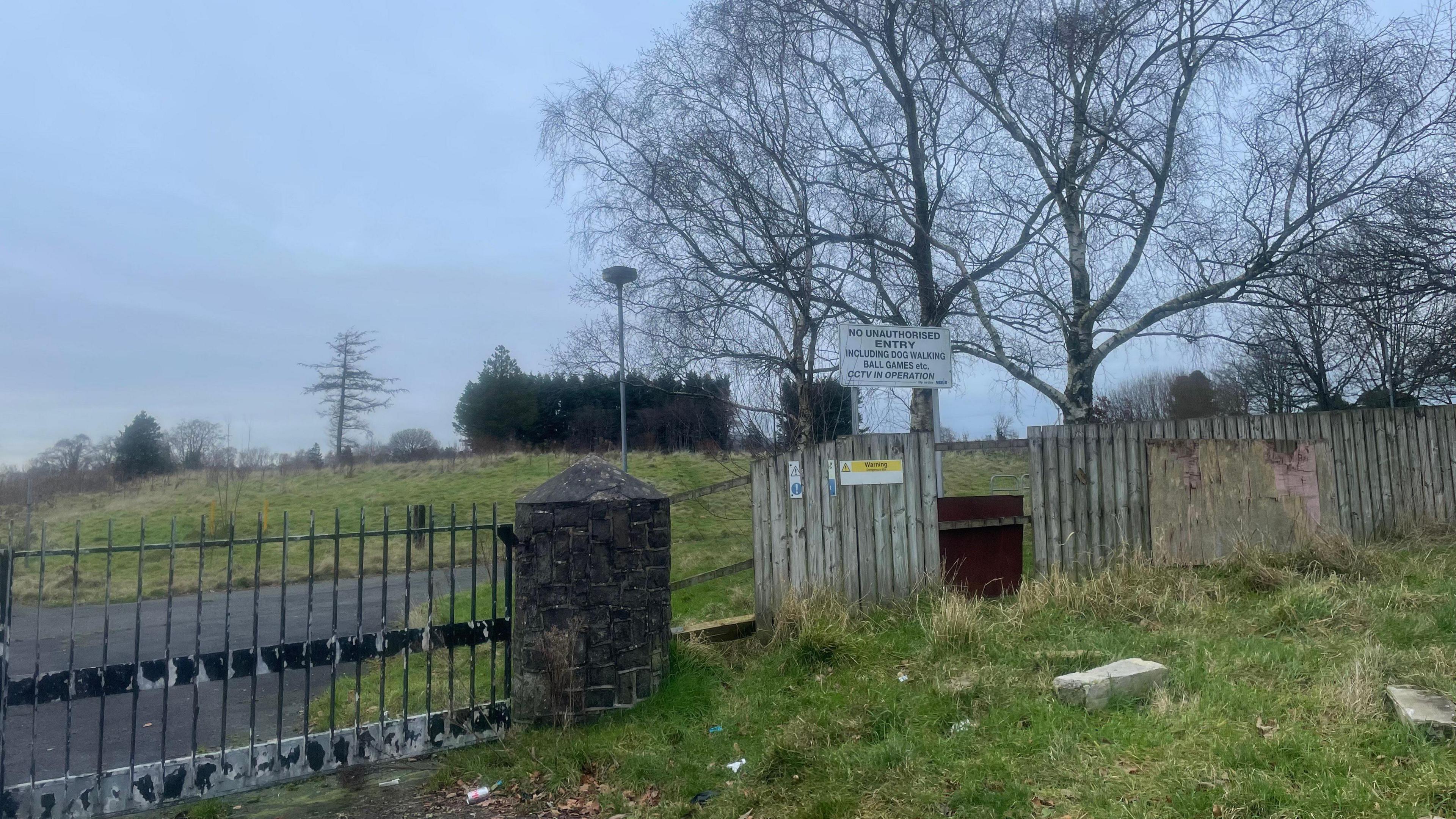 A worn black gate sits attached to a small back stone pillar. Its sits on a tarmac path with grass either side. To its right is a grey fence with gaps in it. Leafless tree's sit behind the fence.