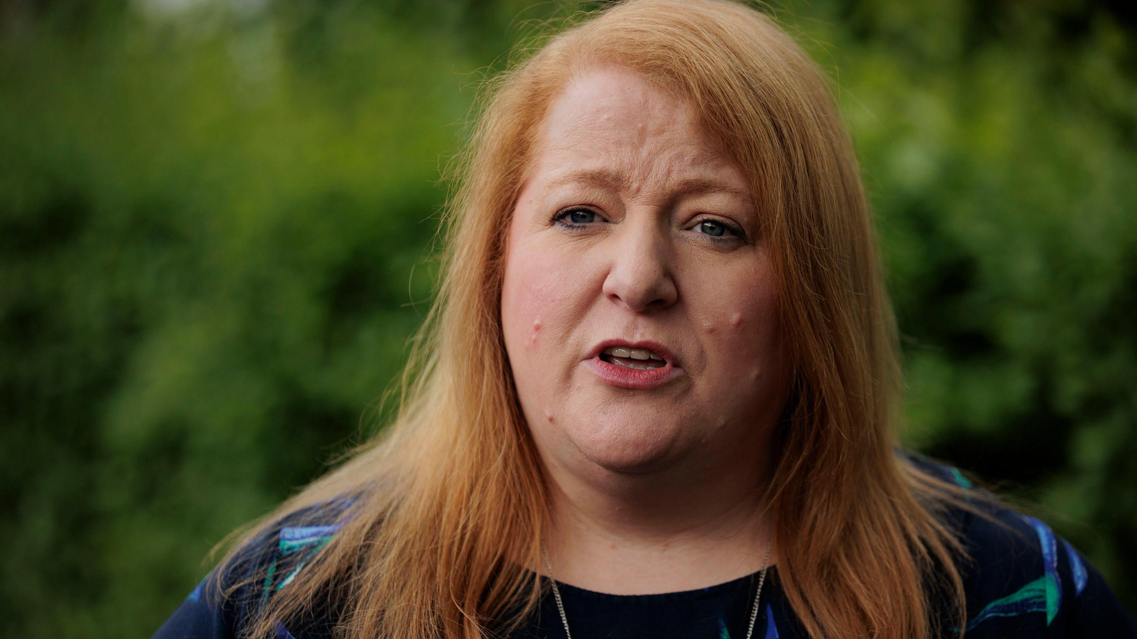 Naomi Long outside with trees behind her. She wears a blue top and has long red hair.