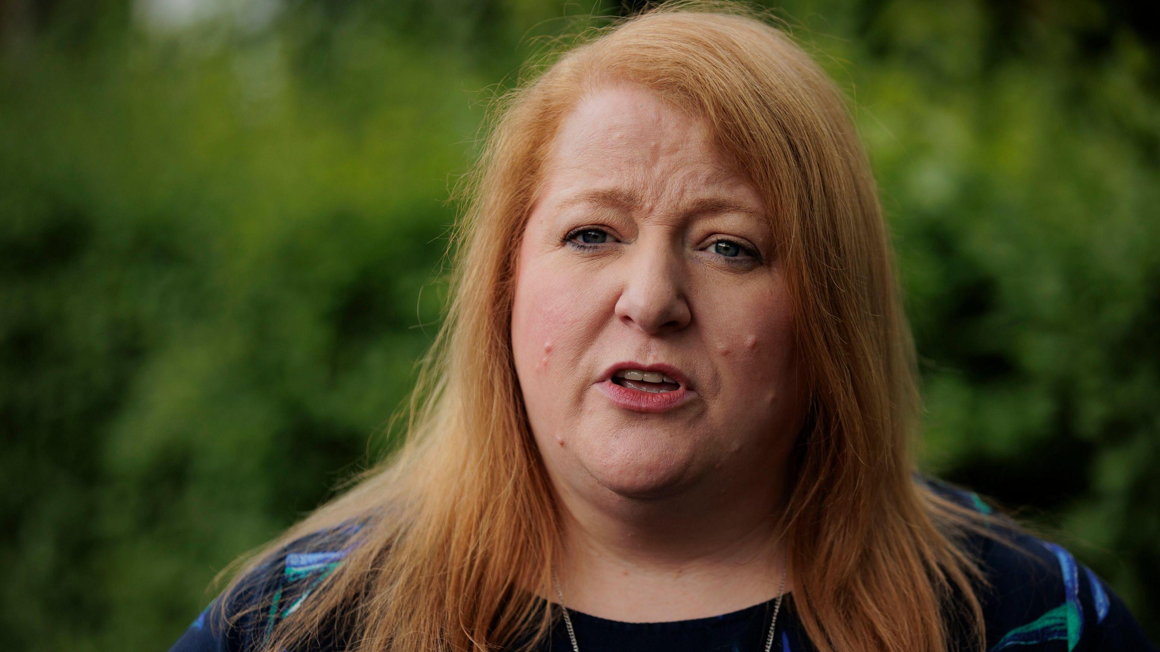 Headshot of Naomi Long speaking outside with some bushes in the background. She is wearing a navy, blue and green top.