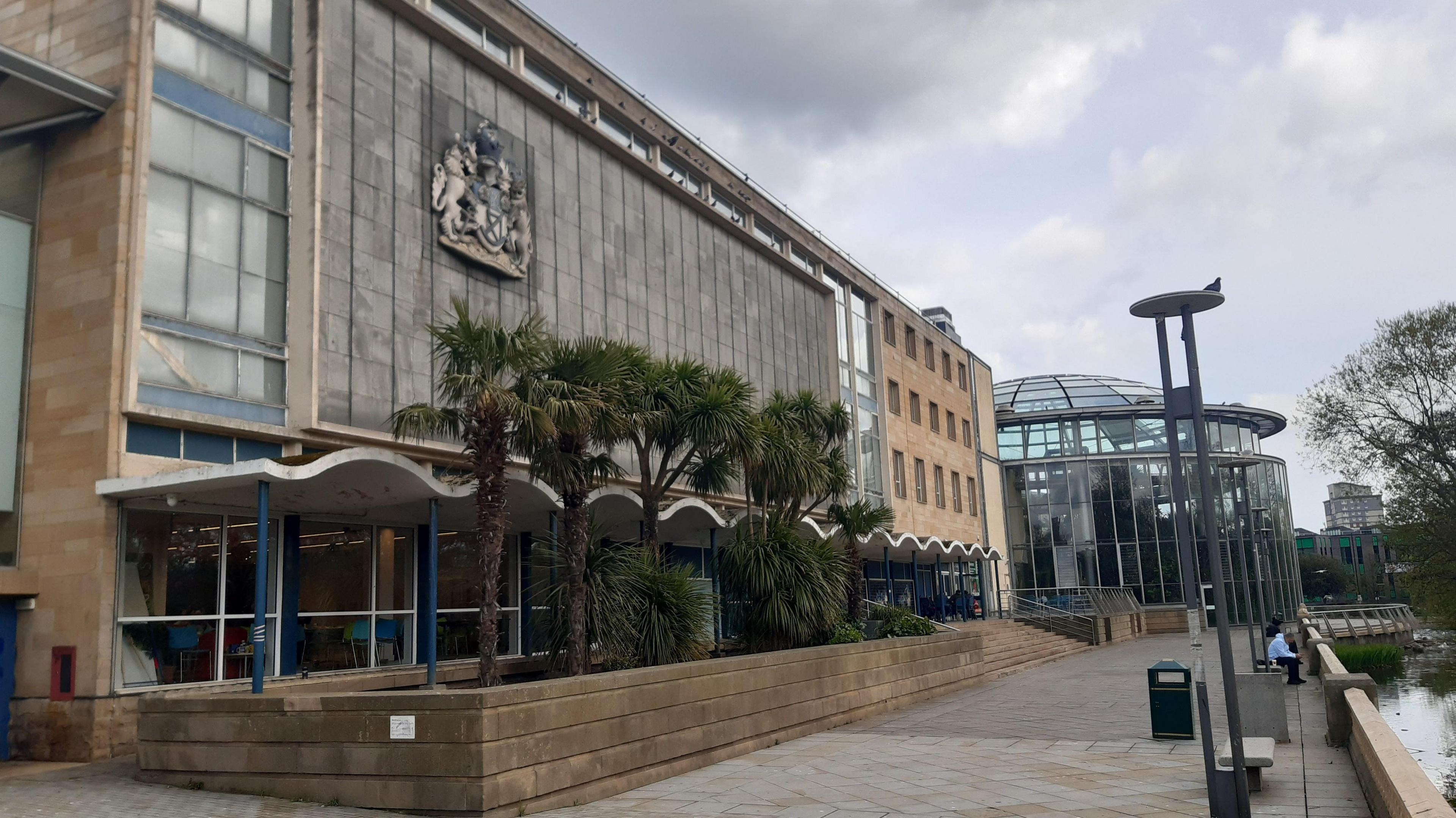 The grey, multi-storey building of Sunderland Museum and Winter Gardens. Several palms are growing in front of it with a pond across a pedestrian area. The cupola glass building of the Winter Gardens can be seen in the distance.