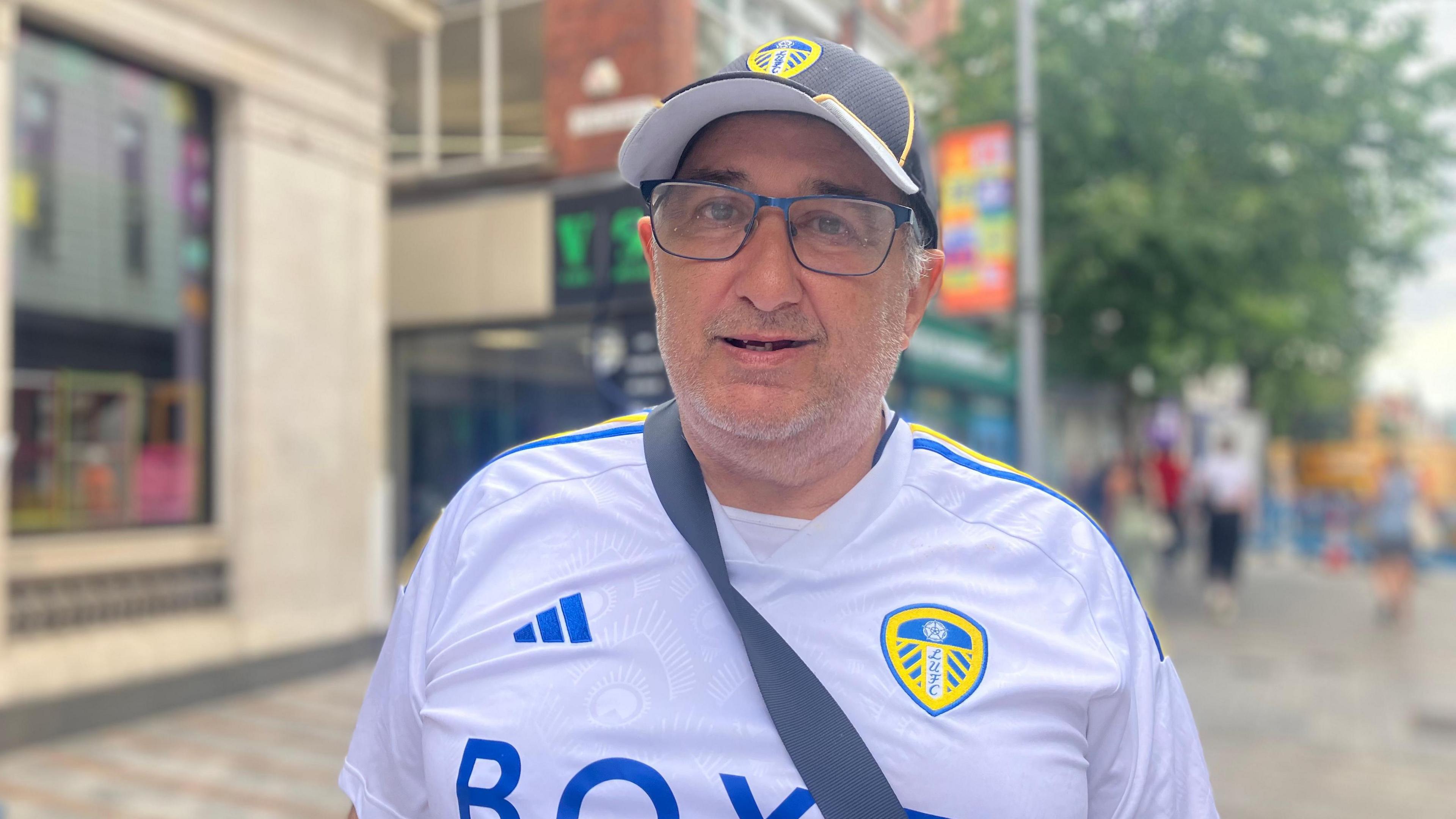 A man with glasses wears a Leeds United Football Club cap and shirt. The strap of a bag crosses his chest. Behind him, Leeds City Centre is blurred.