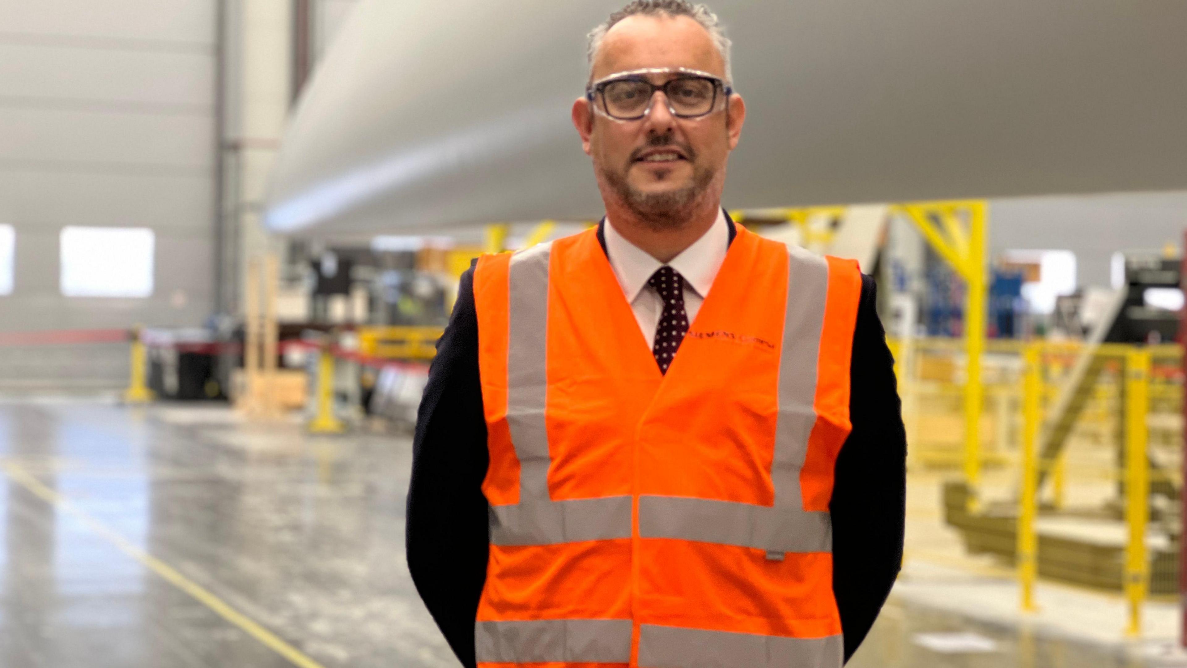 Mr Davidson wearing goggles and an orange and grey hi-vis vest. He is standing on the factory floor with a turbine blade in the background.