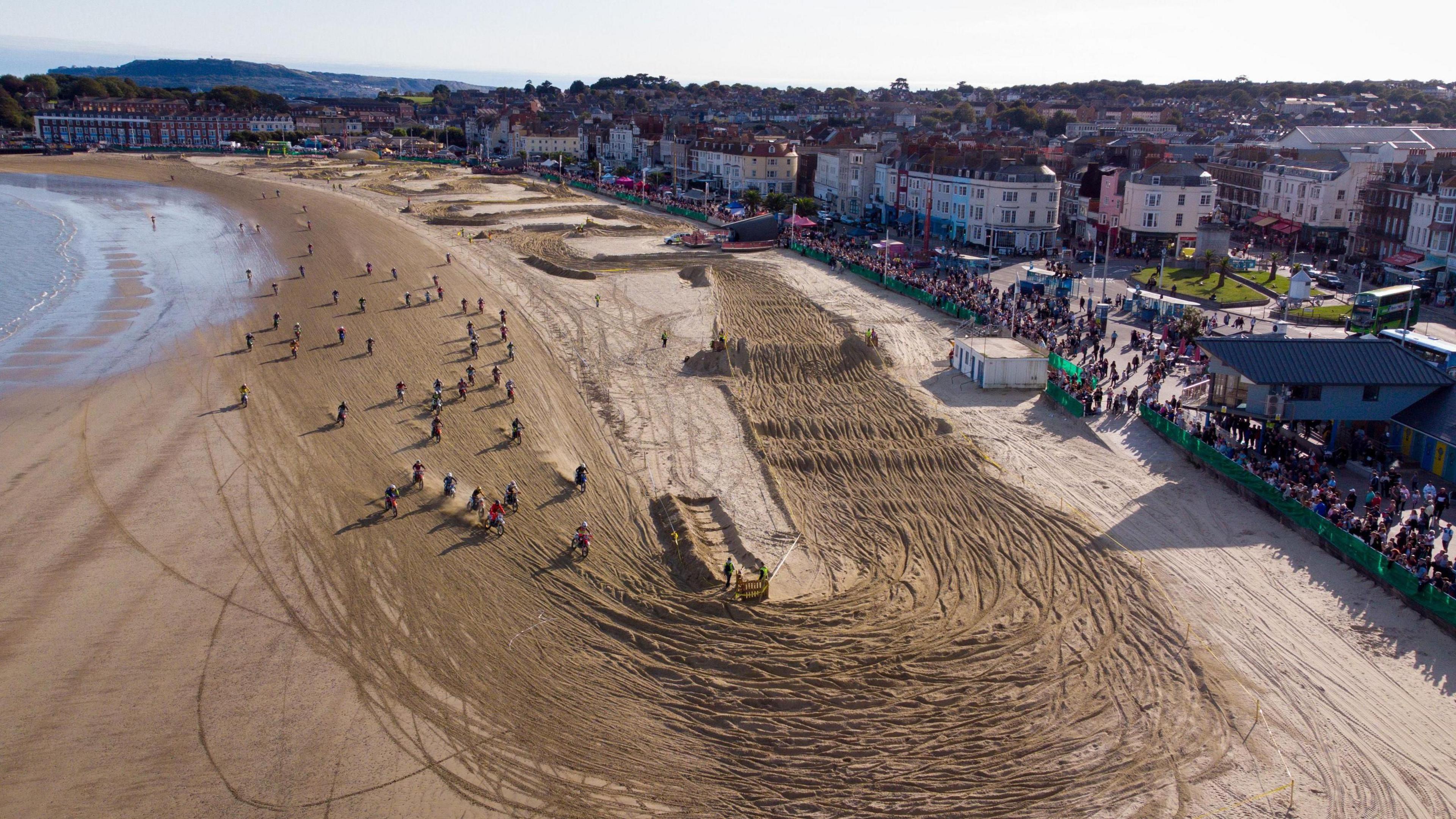 Spectators line the edge of a beach. Its sands have been carved into a race course with mounds and tight turns. Dozens of riders on bikes can be seen to the left of the course on the edge of the sea.
