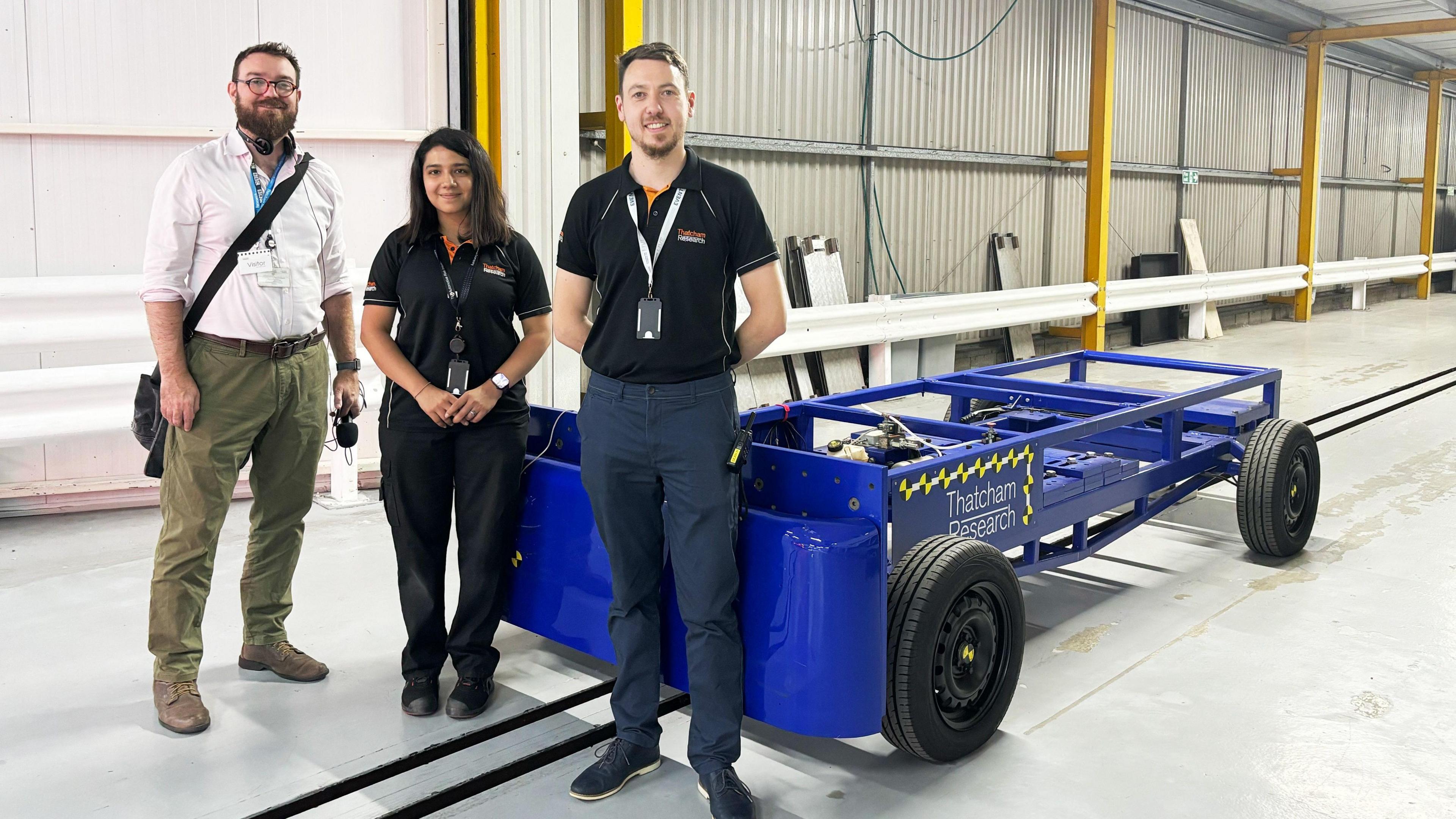 Phil Mercer, wearing a white shirt and green chino trousers, stood next to Mitali Shah and Sean Hoad, who are both wearing blak polo shirts and trousers. They are all stood in front of a blue trolley.