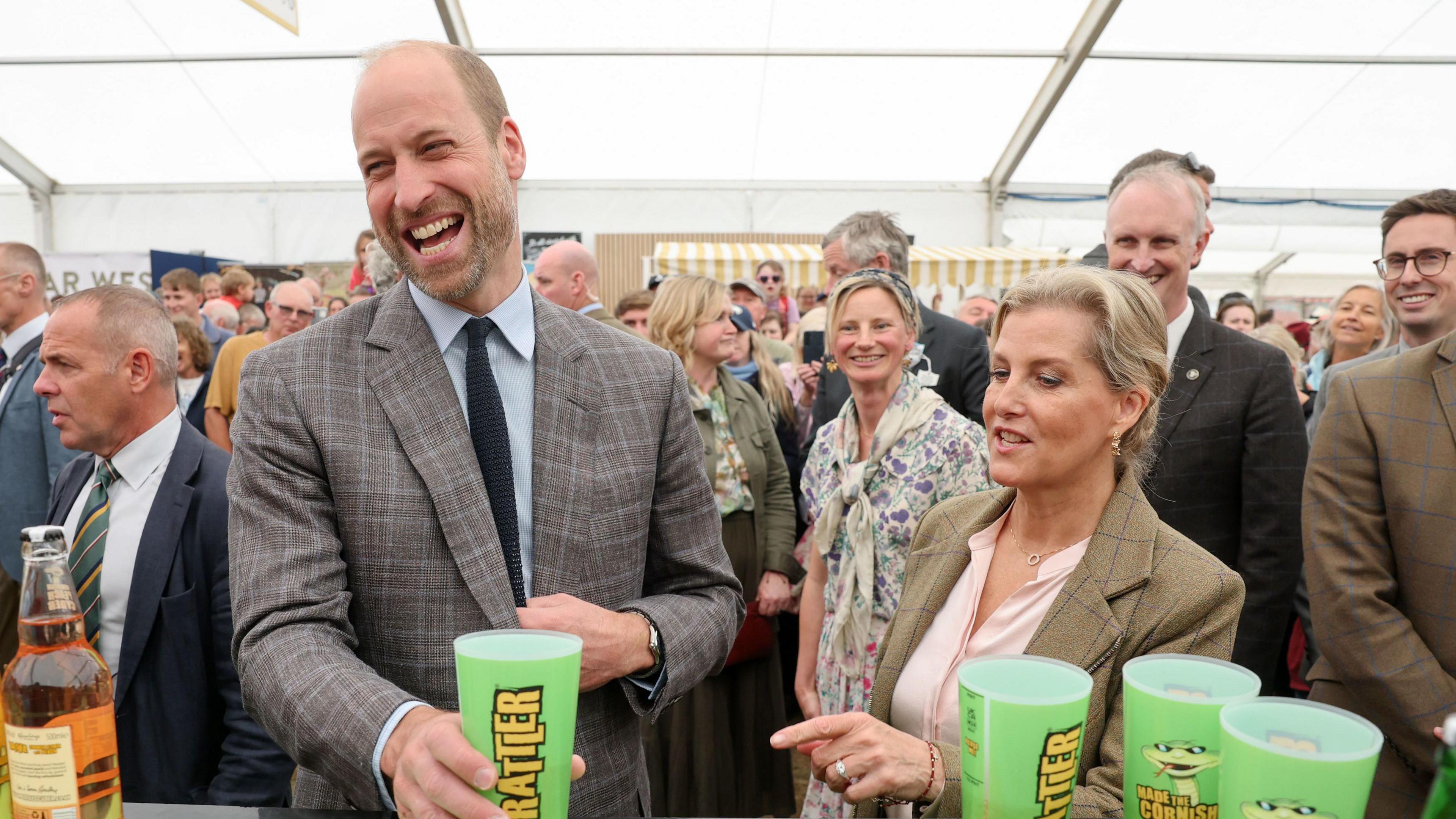 The Prince of Wales, known as the Duke of Cornwall while in Cornwall, and Sophie the Duchess of Edinburgh try local cider during a visit to the Food and Farming Pavilion during the Royal Cornwall Show. They are stood in a tent with people around them. Prince William is holding a green Rattler cider cup. 
