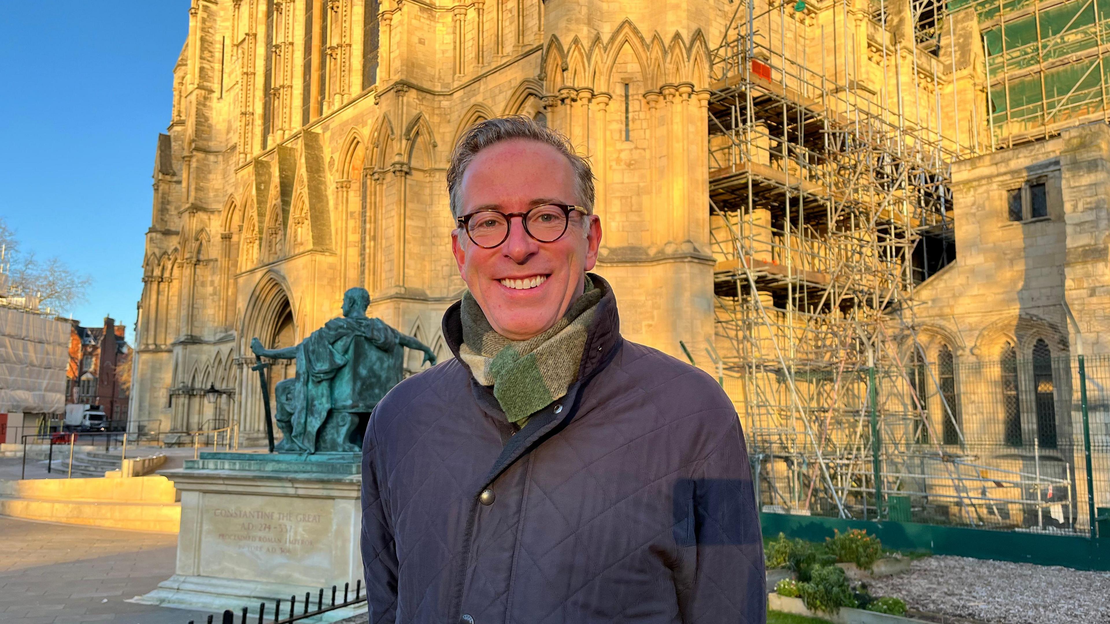 A man in a blue coat with a green scarf is standing in front of York Minster with a bronze statue of Constantine the Great behind his right shoulder.