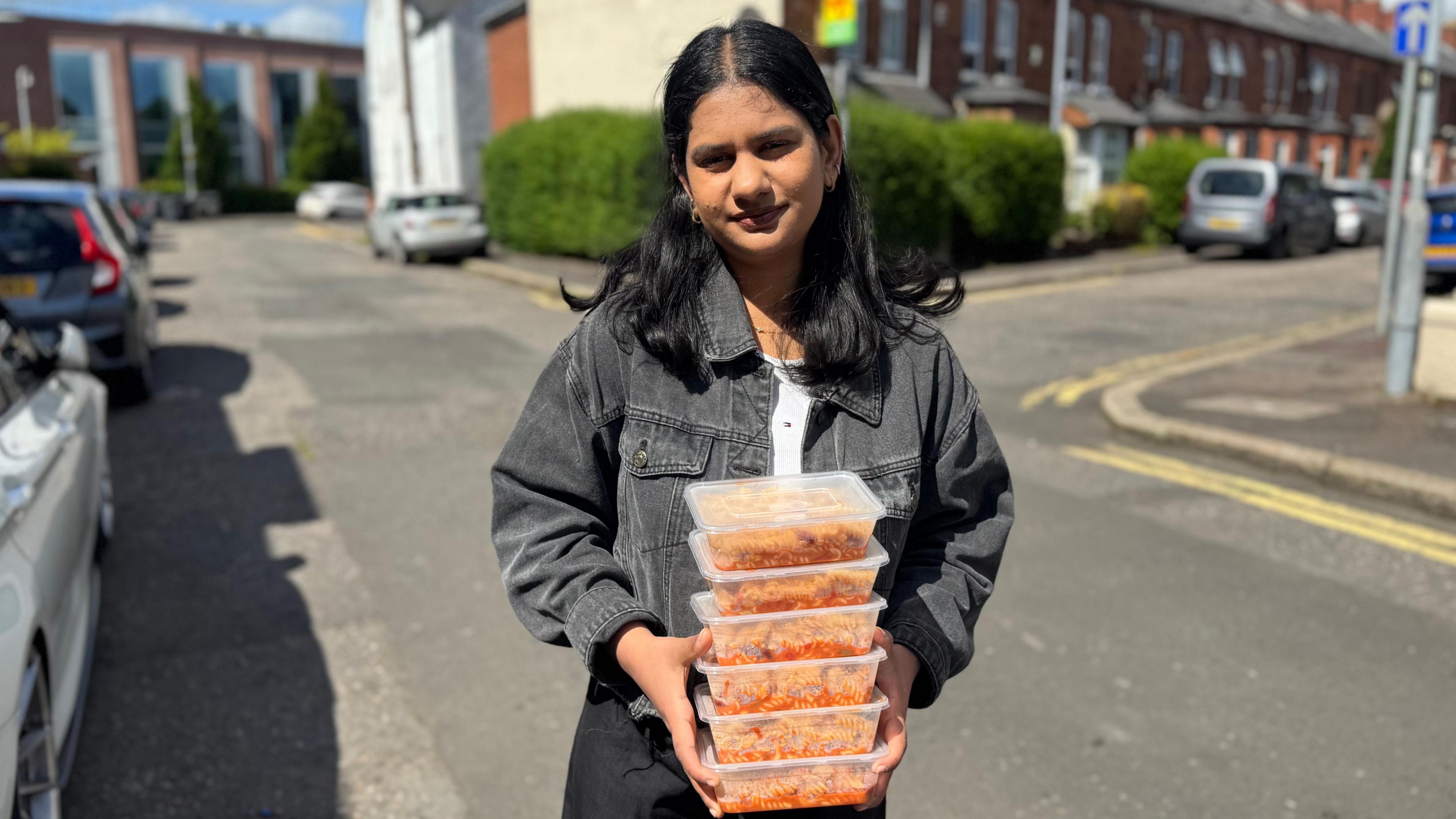 A young indian woman is standing in a residential street. She is wearing black trousers, a black denim jacket and a white t-shirt. She has black hair. She is holding six clear boxes of tomato pasta. 