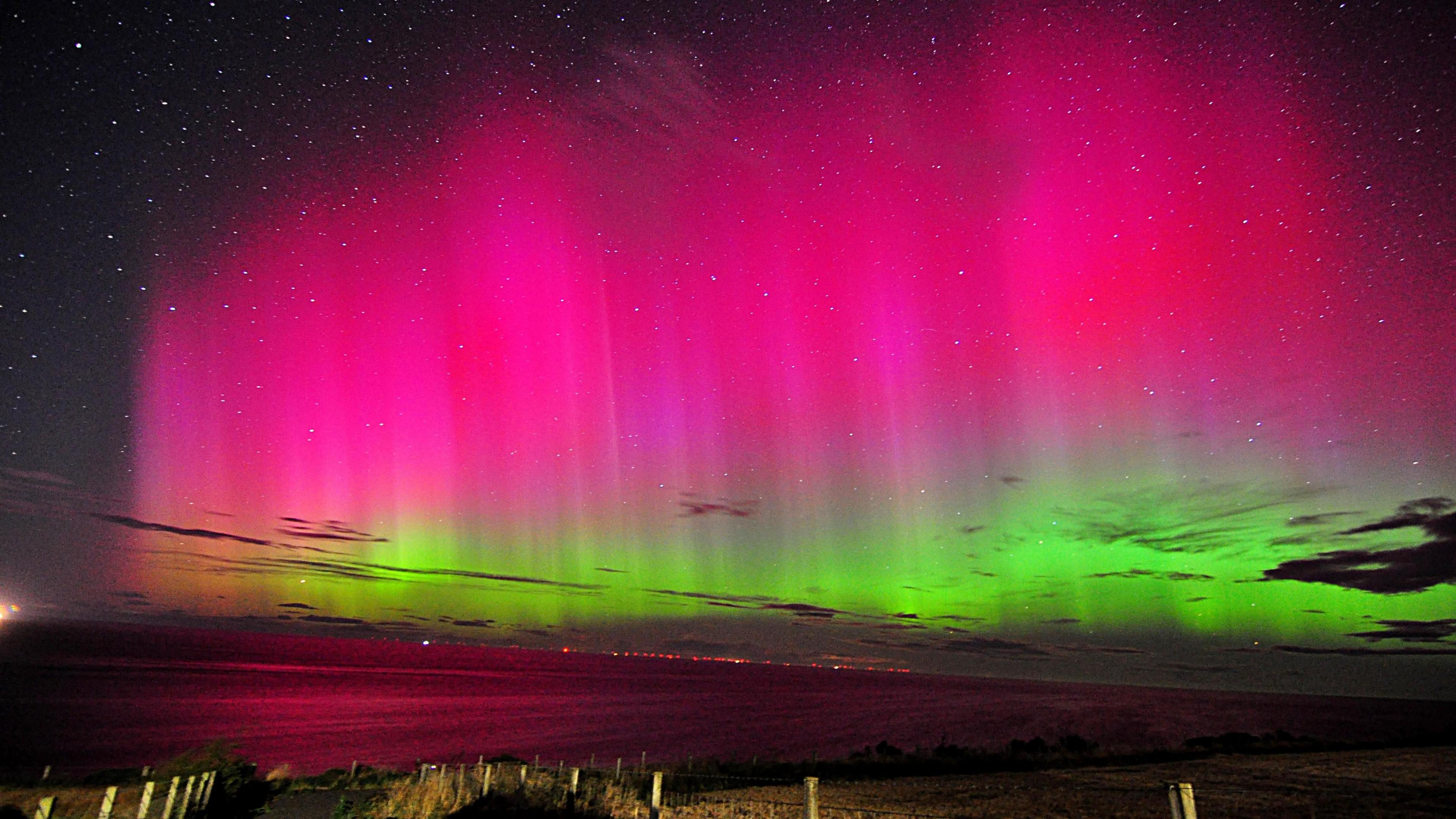 A picture of the night sky across a big empty expanse. The sky is lit up bright green and pink in waves, showing the aurora, in front of a sparkling night sky filled with stars