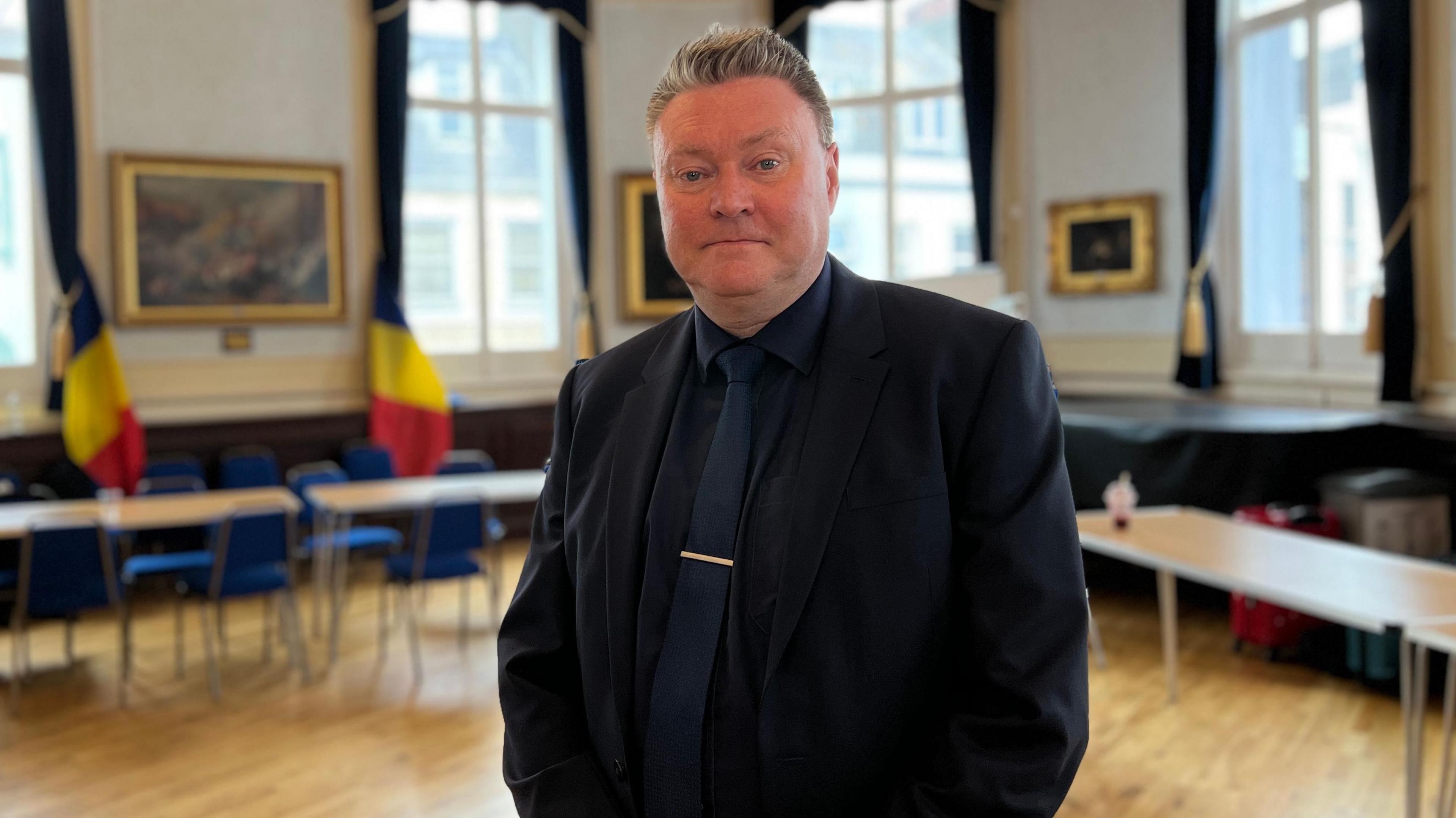 Deputy Rob Ward looks at the camera and there are desks and chairs behind him as well as two Romanian flags