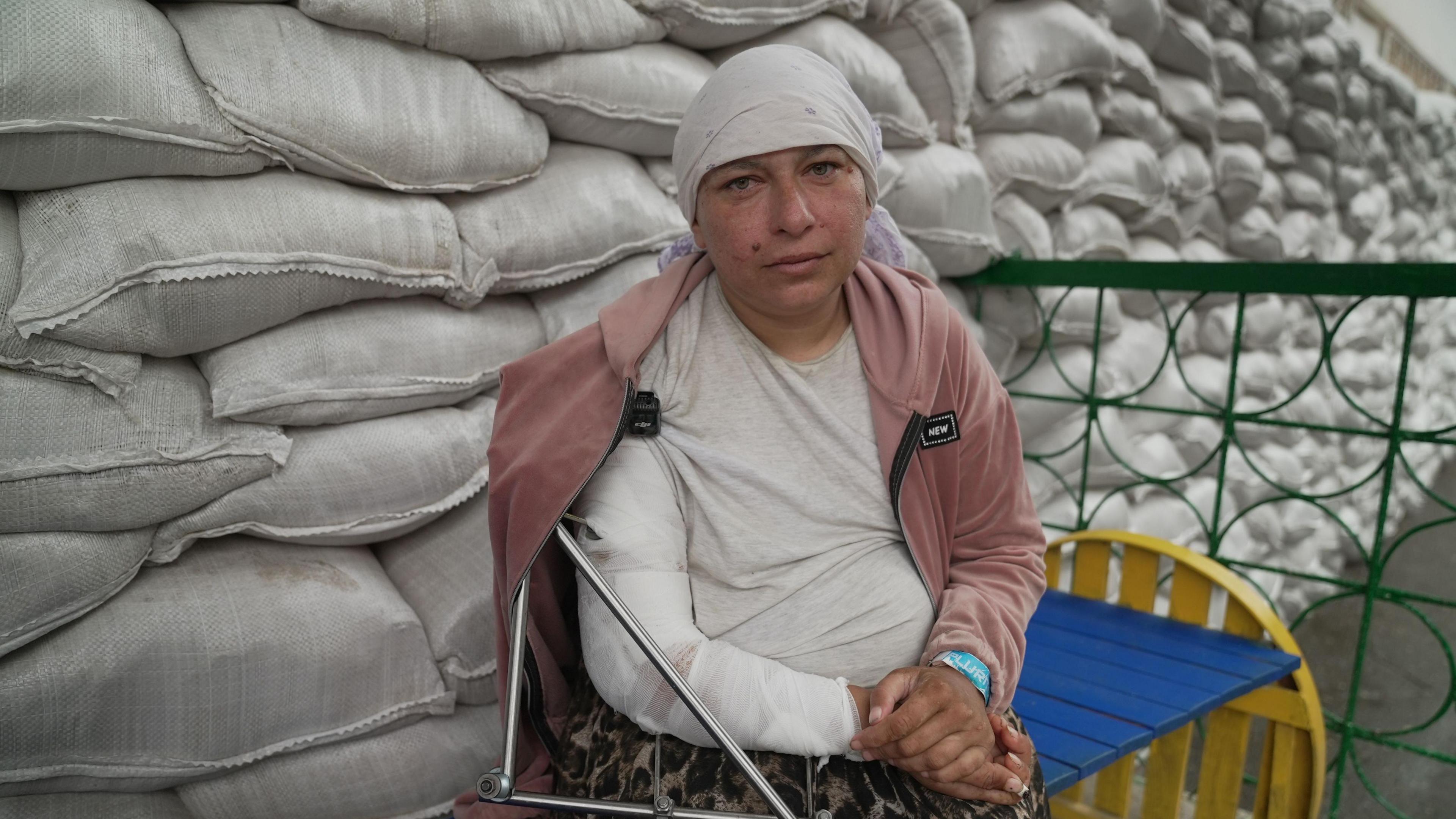A woman wearing a white headcover with her arm in a sling sits on a bench